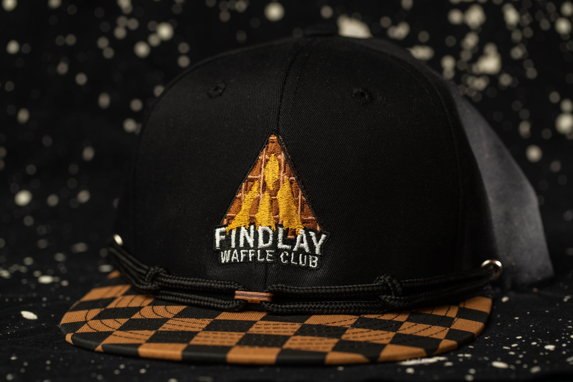 A black baseball cap with a checkered pattern and a logo on it, against a dark background with white speckles.