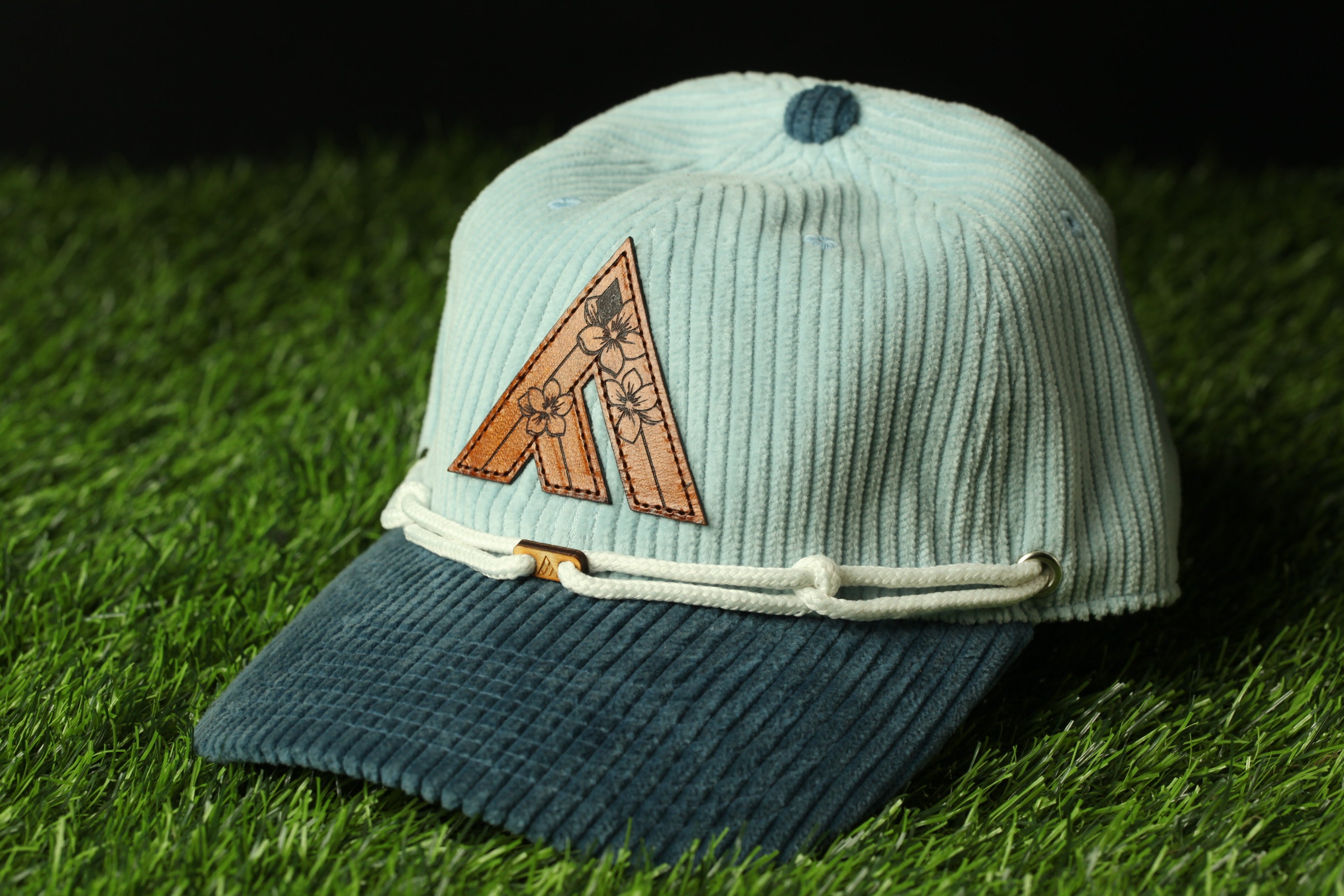 A light blue baseball cap with a brown floral logo on the front, resting on a grassy surface.