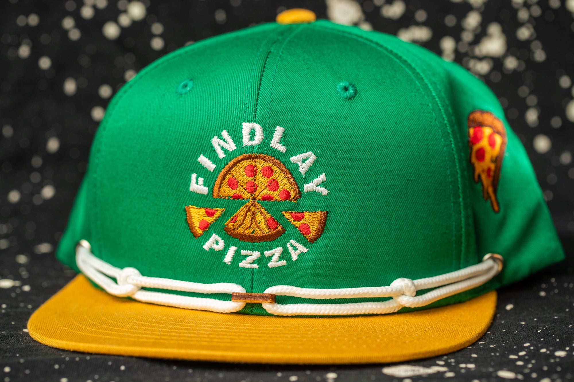 A green baseball cap with a yellow brim and white stitching, featuring a logo for Findlay Pizza.