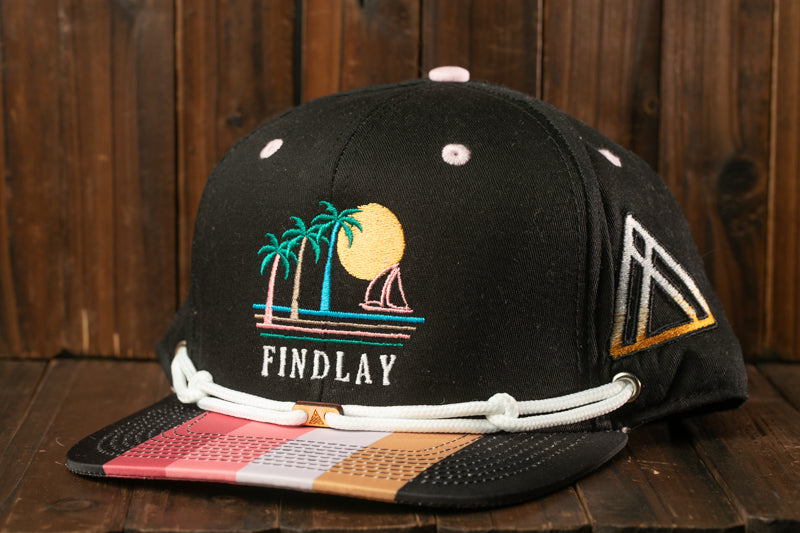 A black baseball cap with a colorful graphic design on the front, featuring palm trees, a sailboat, and a sunset.