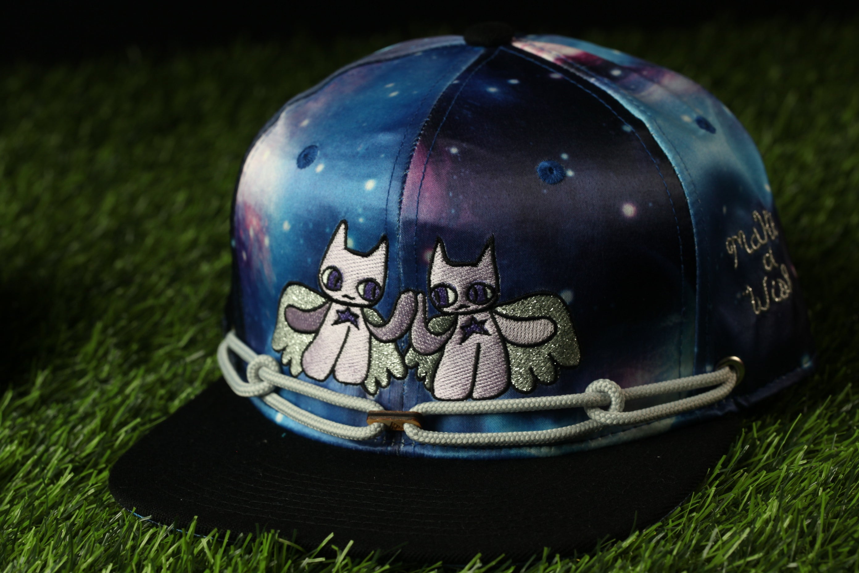 A colorful baseball cap with a galaxy-themed design, featuring two cat-like figures with wings.