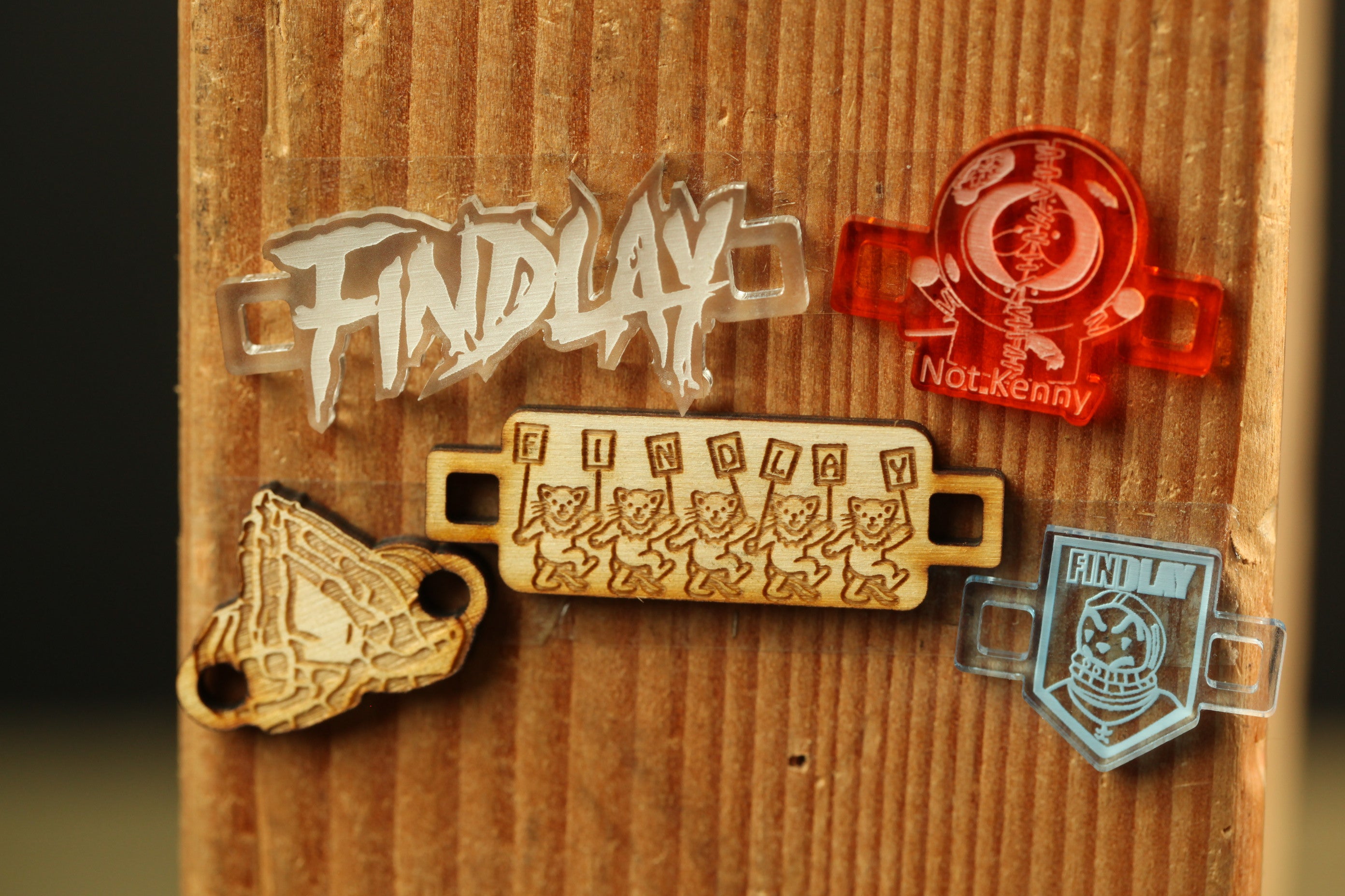 a wooden surface with various objects attached to it, including a clear plastic object with the word "FINDlay" written on it, a wooden object with a group of cartoon cats, and a red object with the text "NotKenny" written on it.