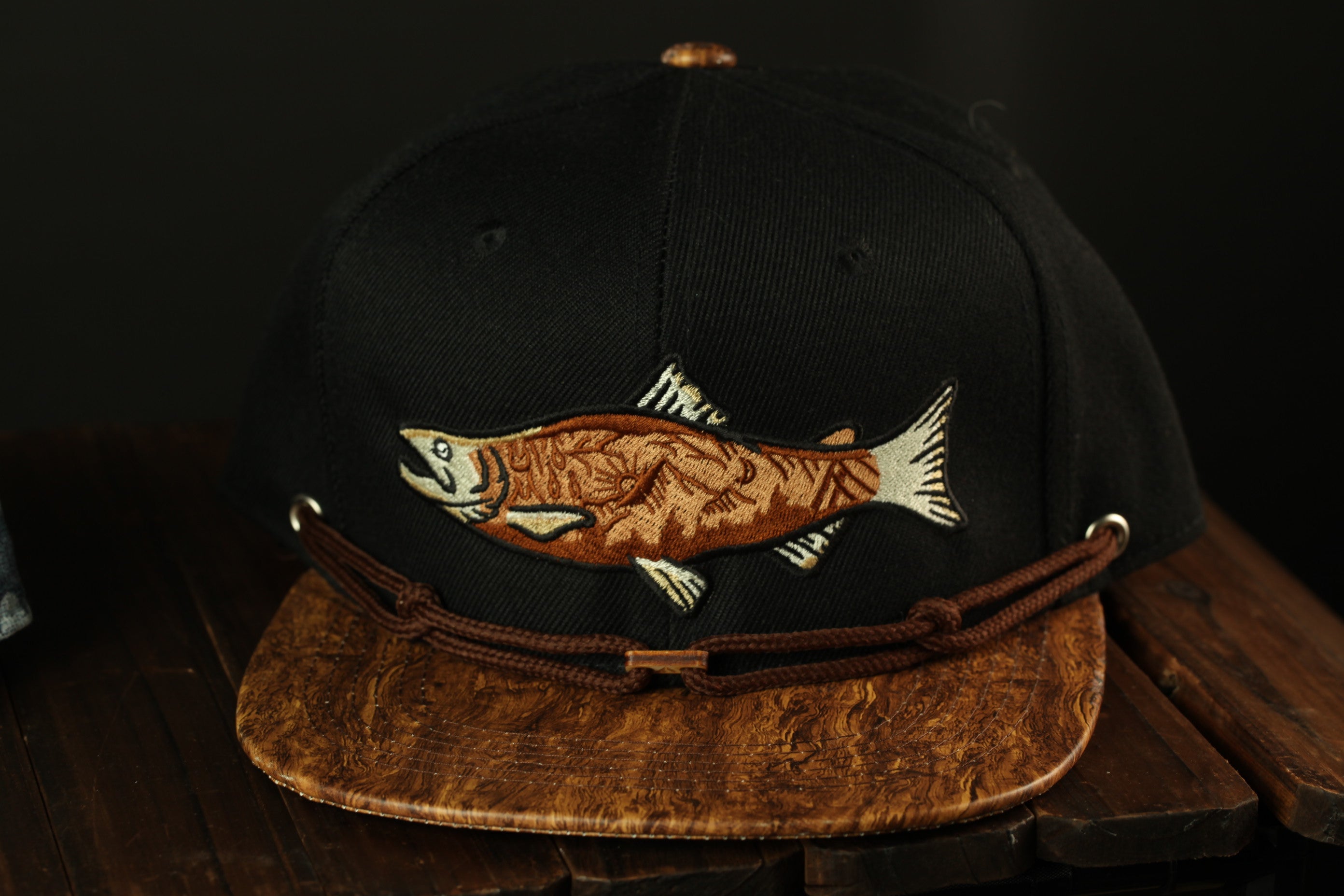A black baseball cap with a brown leather brim and a fish embroidered on the front, sitting on a wooden surface.