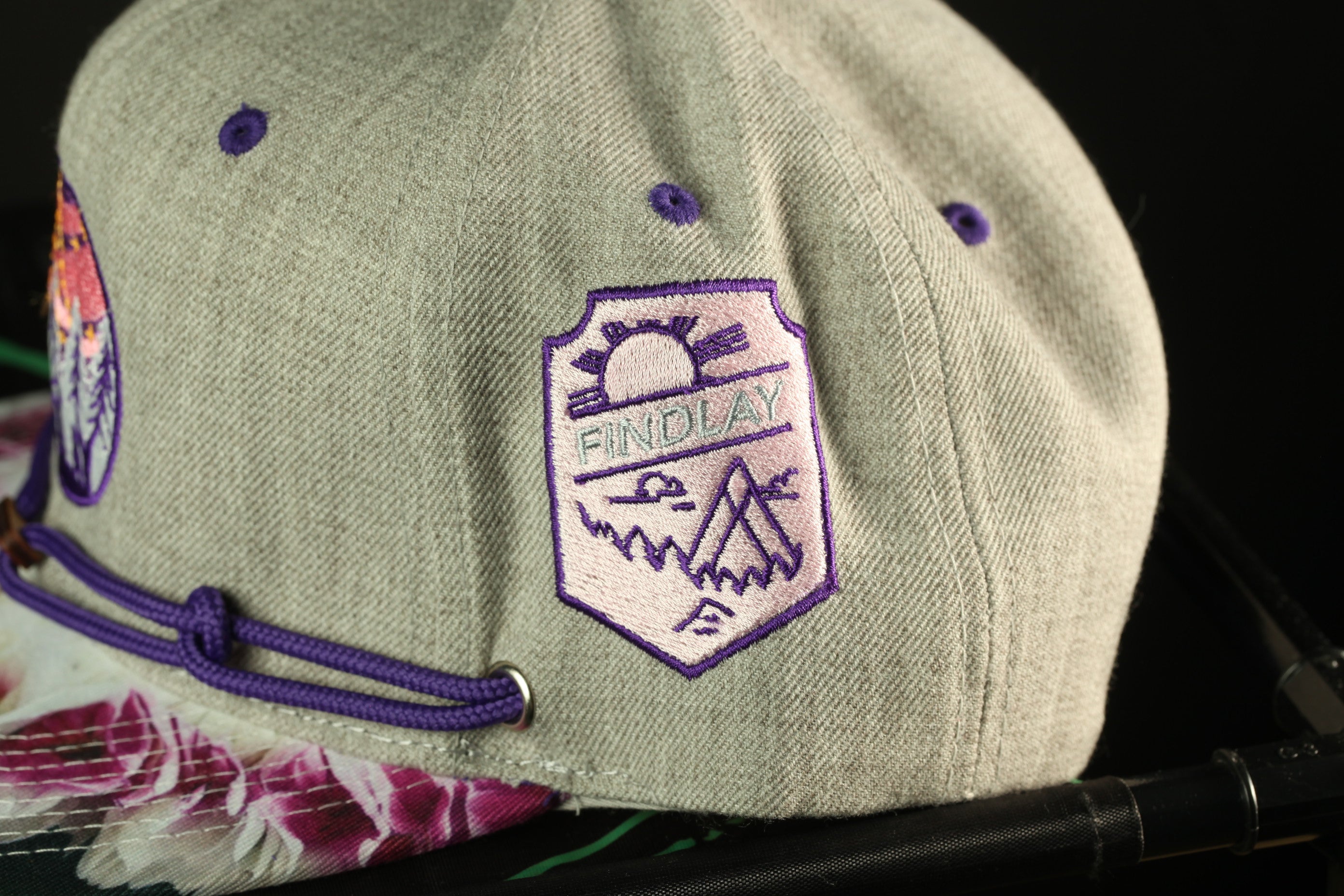 A gray baseball cap with purple embroidered patches and a sun-like symbol on the front.