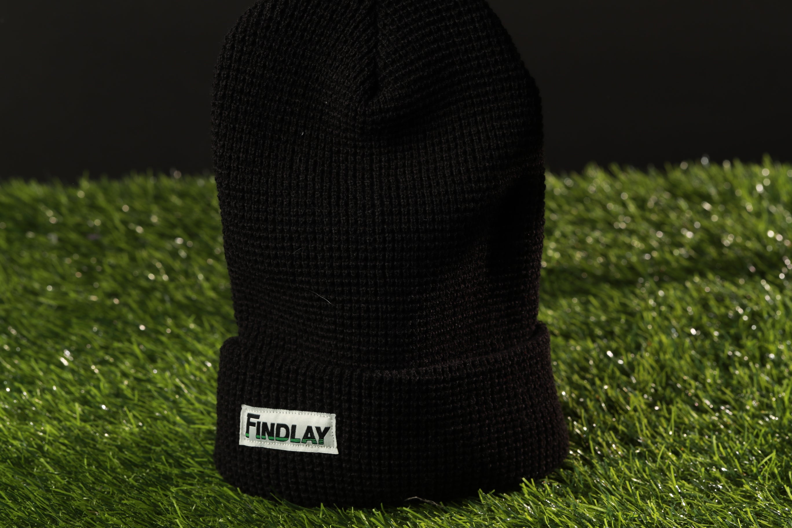 A black knit beanie with a white "Findlay" label is placed on a grassy surface.