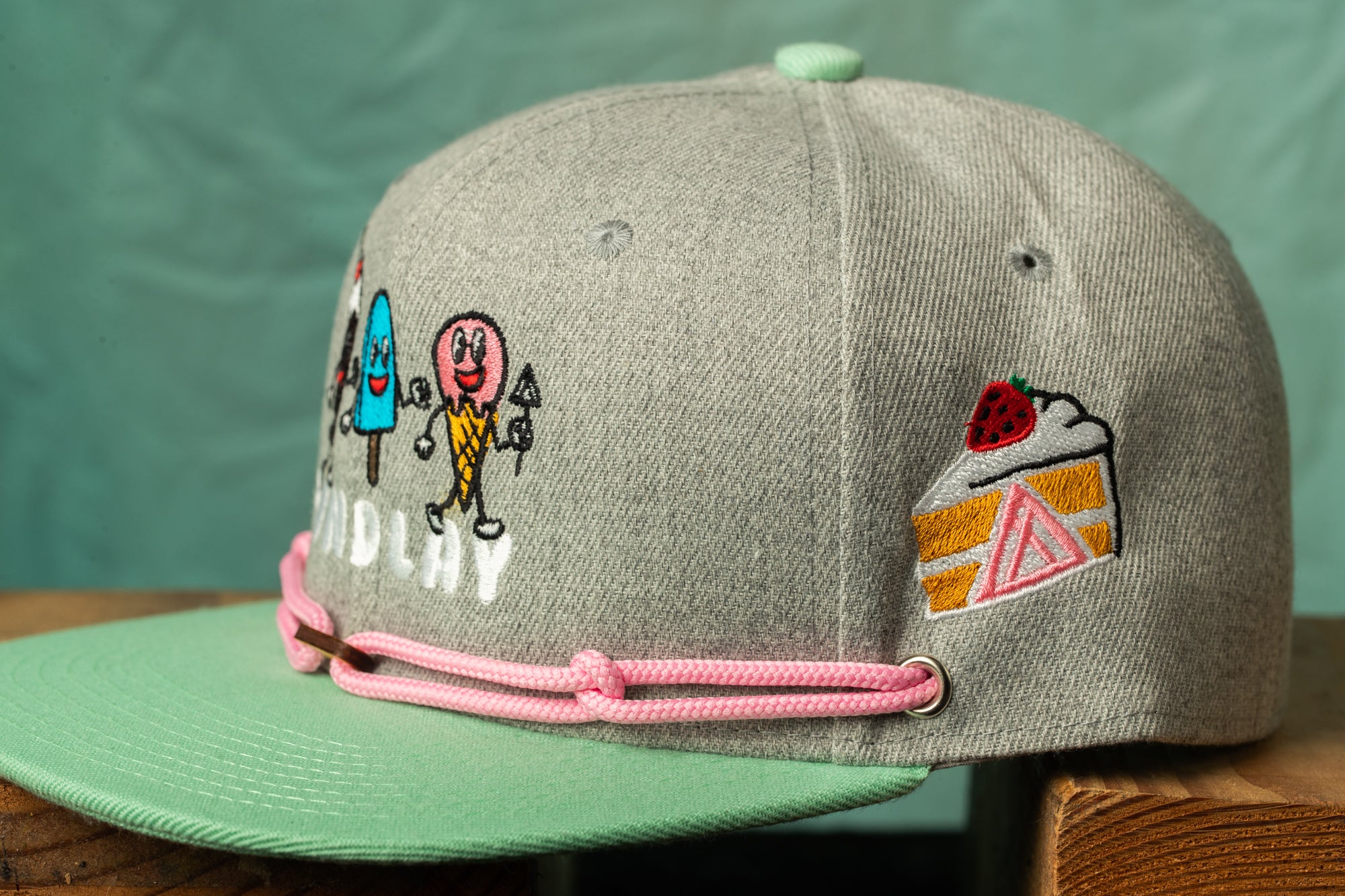A gray baseball cap with colorful cartoon characters and a slice of cake embroidered on it.