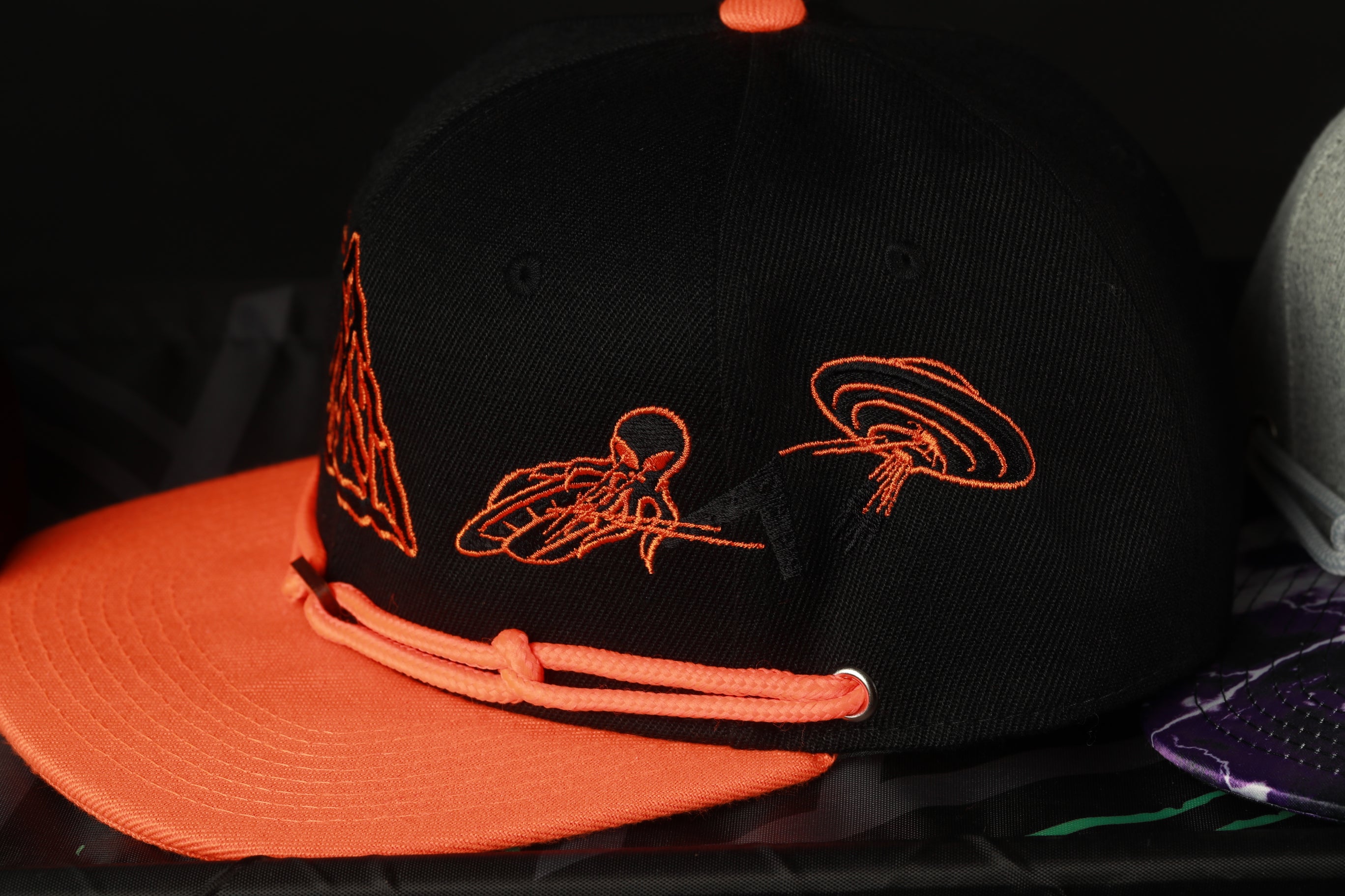 A black baseball cap with orange and black embroidered designs, including a triangle and a spider, is shown in the image.