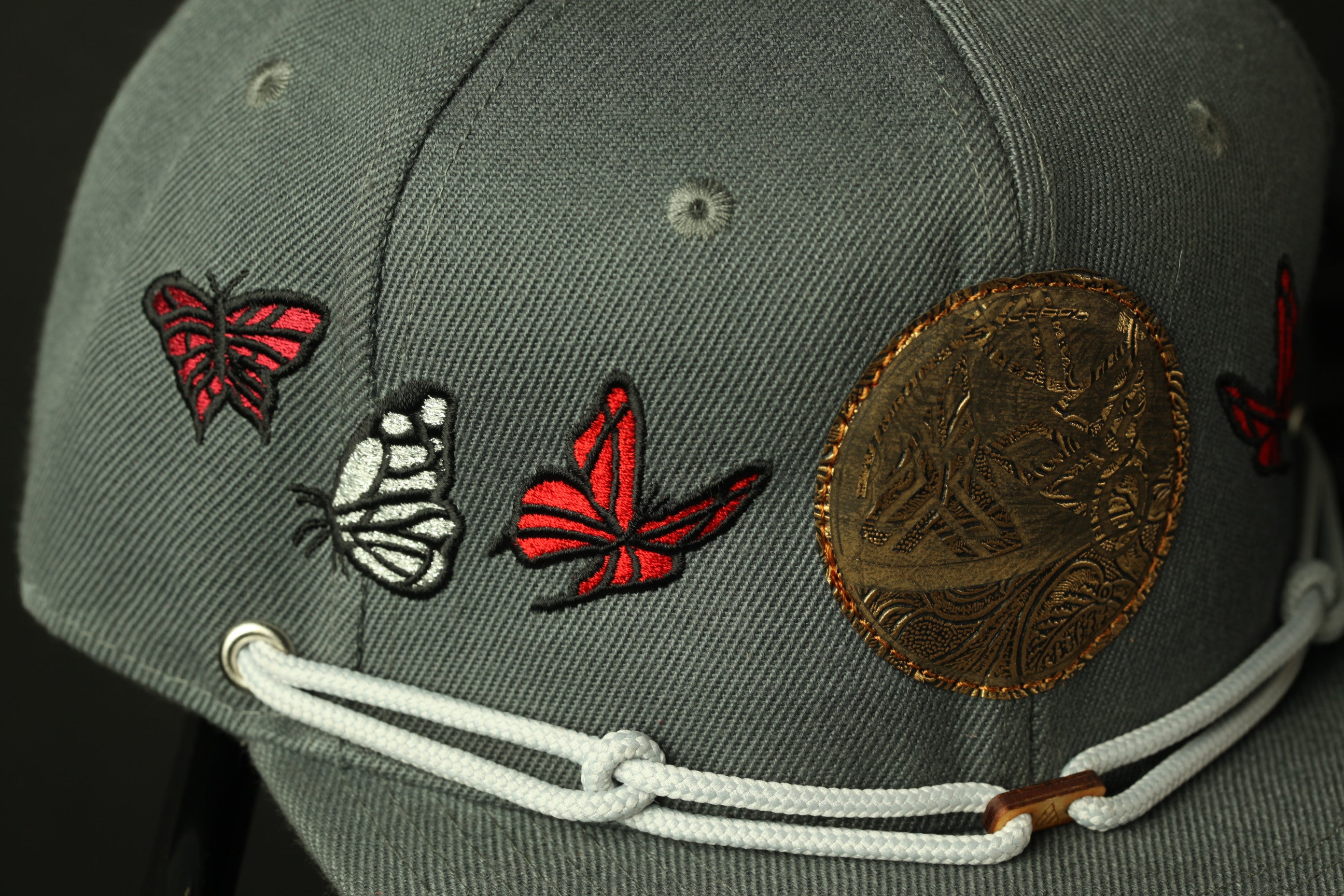 A close-up view of a gray baseball cap with various embroidered designs, including butterflies and a circular emblem.