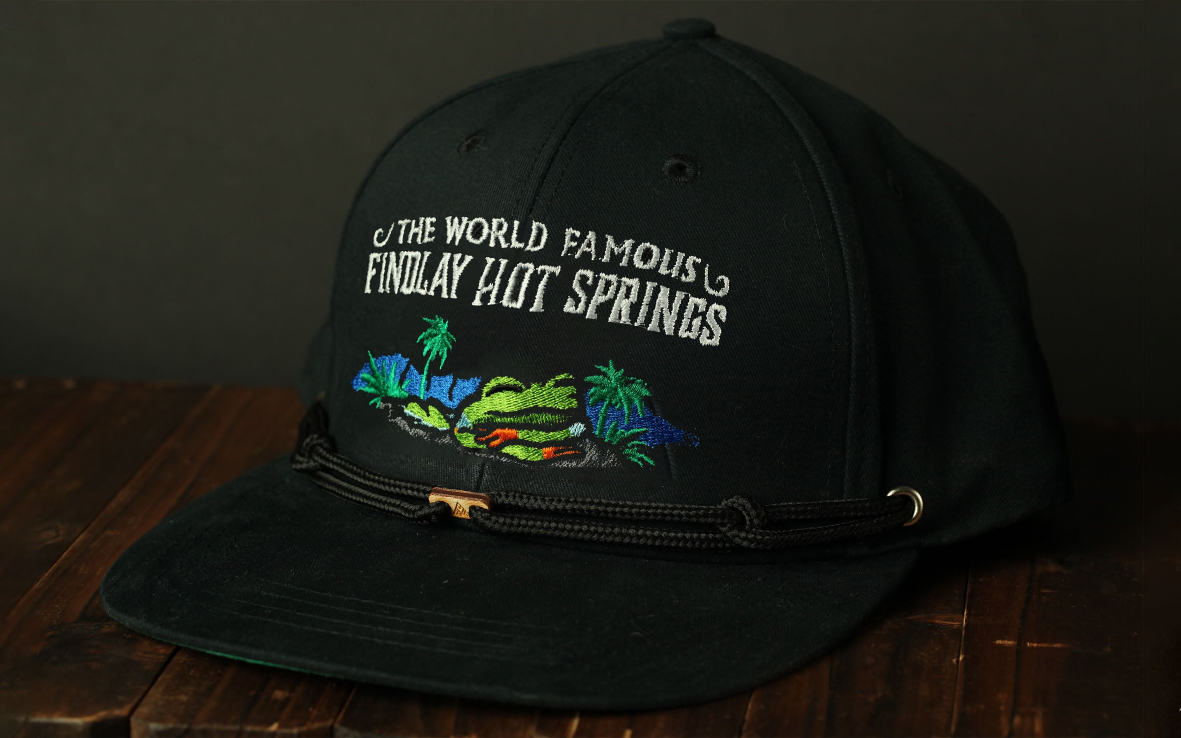 A black baseball cap with a printed design on the front, featuring text and a tropical landscape.