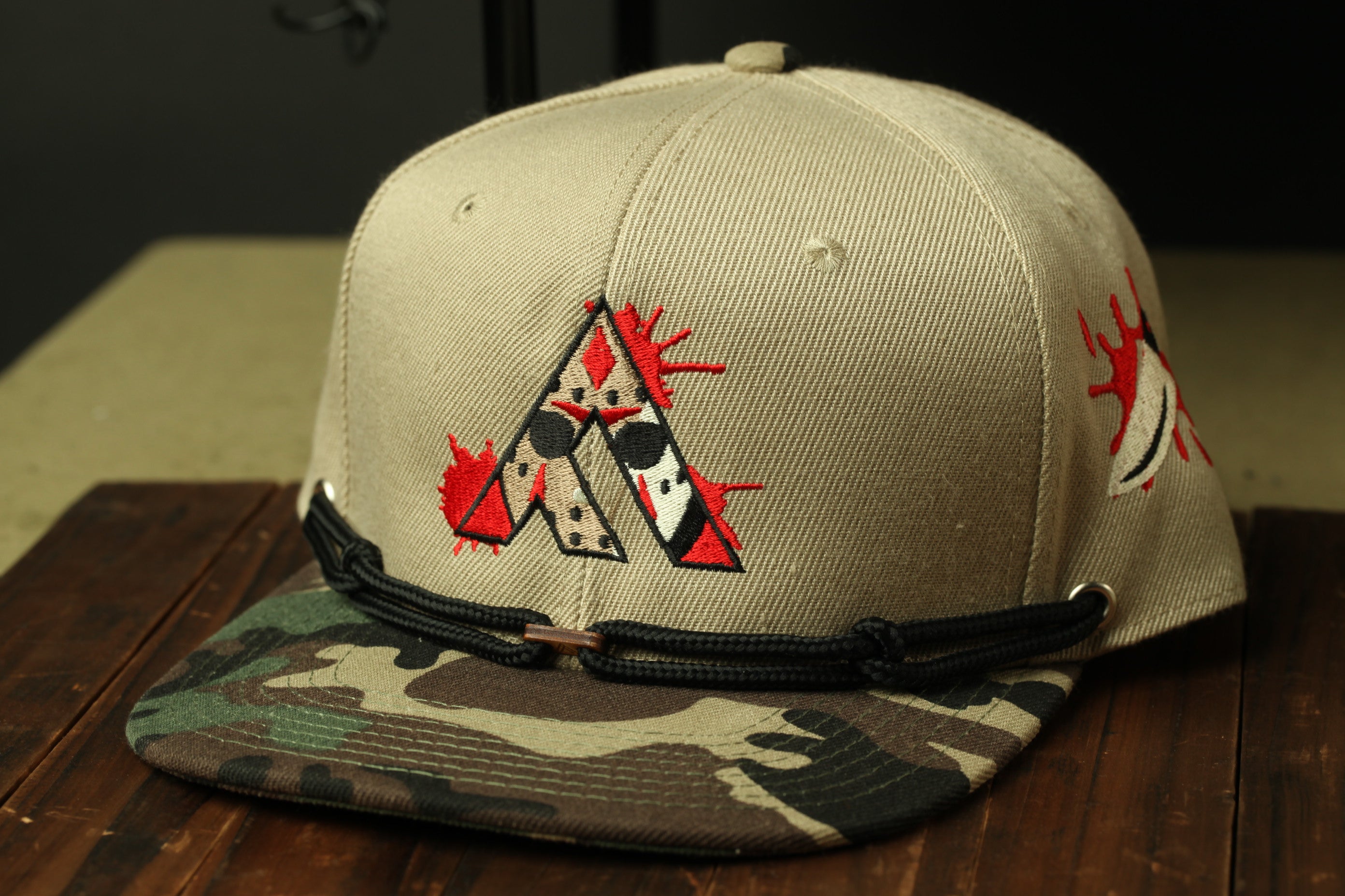 A beige baseball cap with a camouflage pattern and a red and black graphic design on the front.