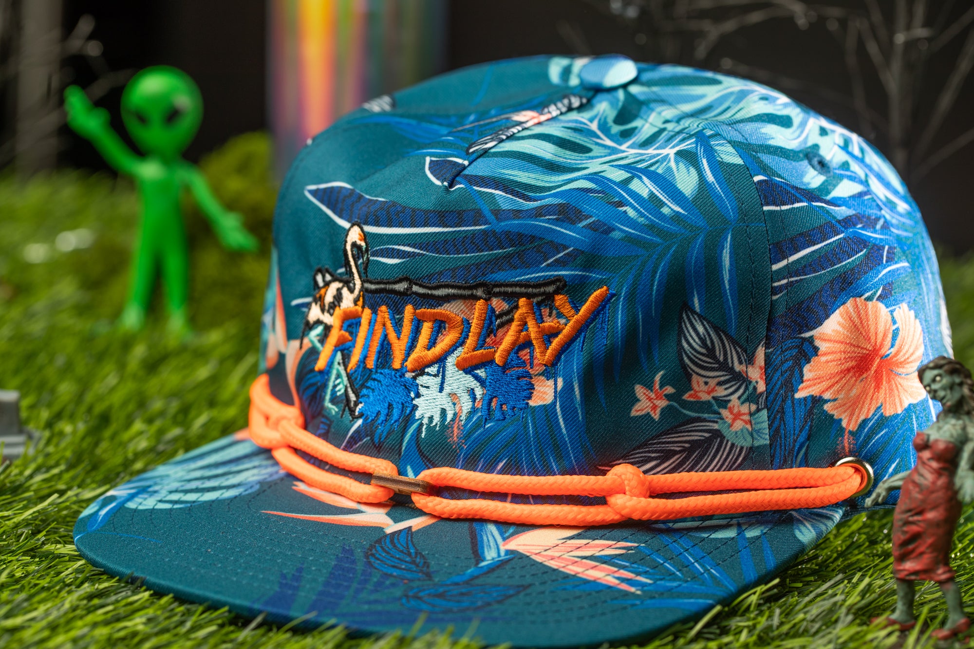 A colorful baseball cap with a tropical print design, featuring the word "FINDlay" embroidered on it. The cap is placed on a grassy surface, with a small green alien figurine and a toy figurine of a person in the background.