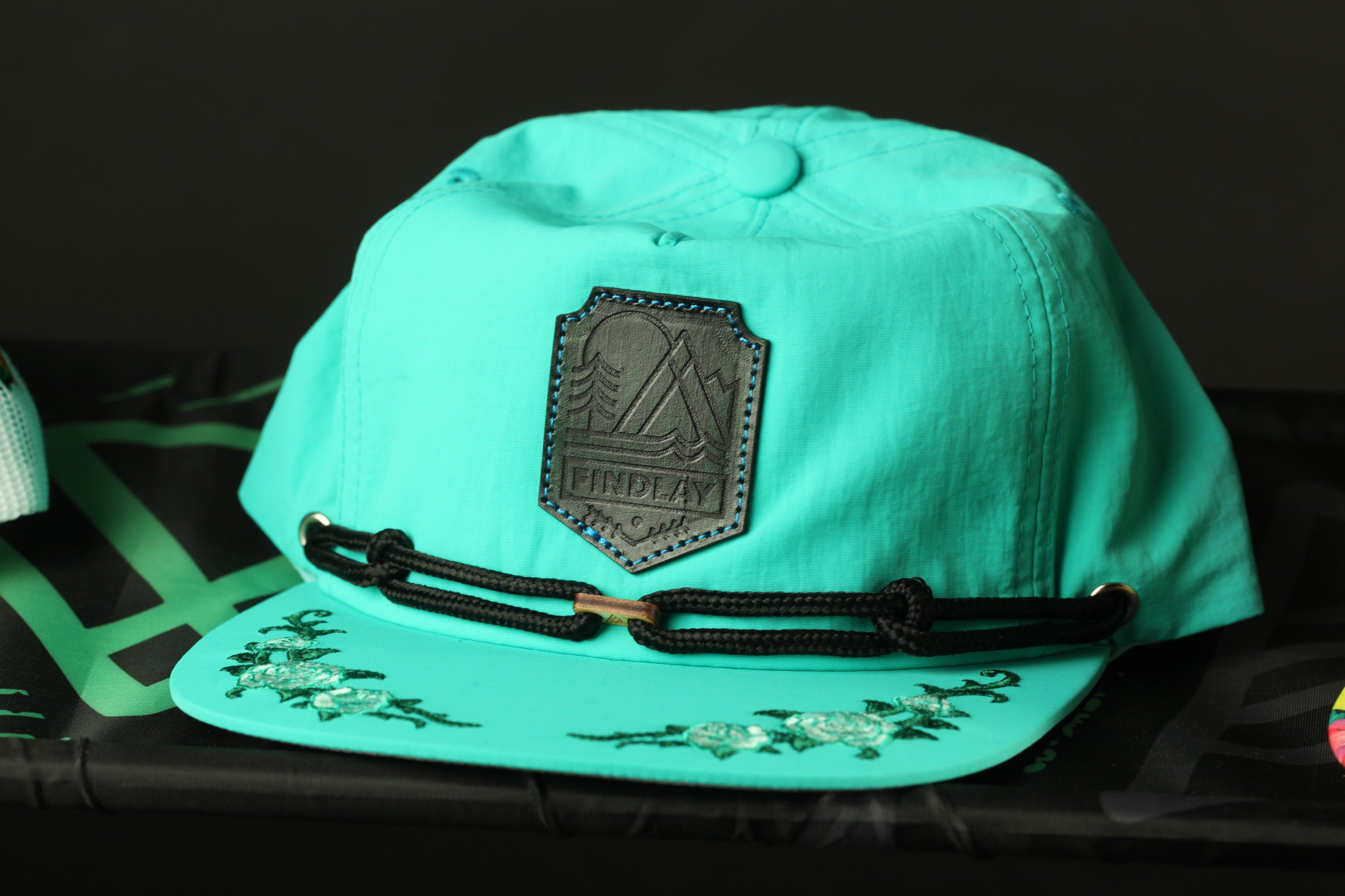 A turquoise baseball cap with a black leather patch and floral embroidery on the brim.