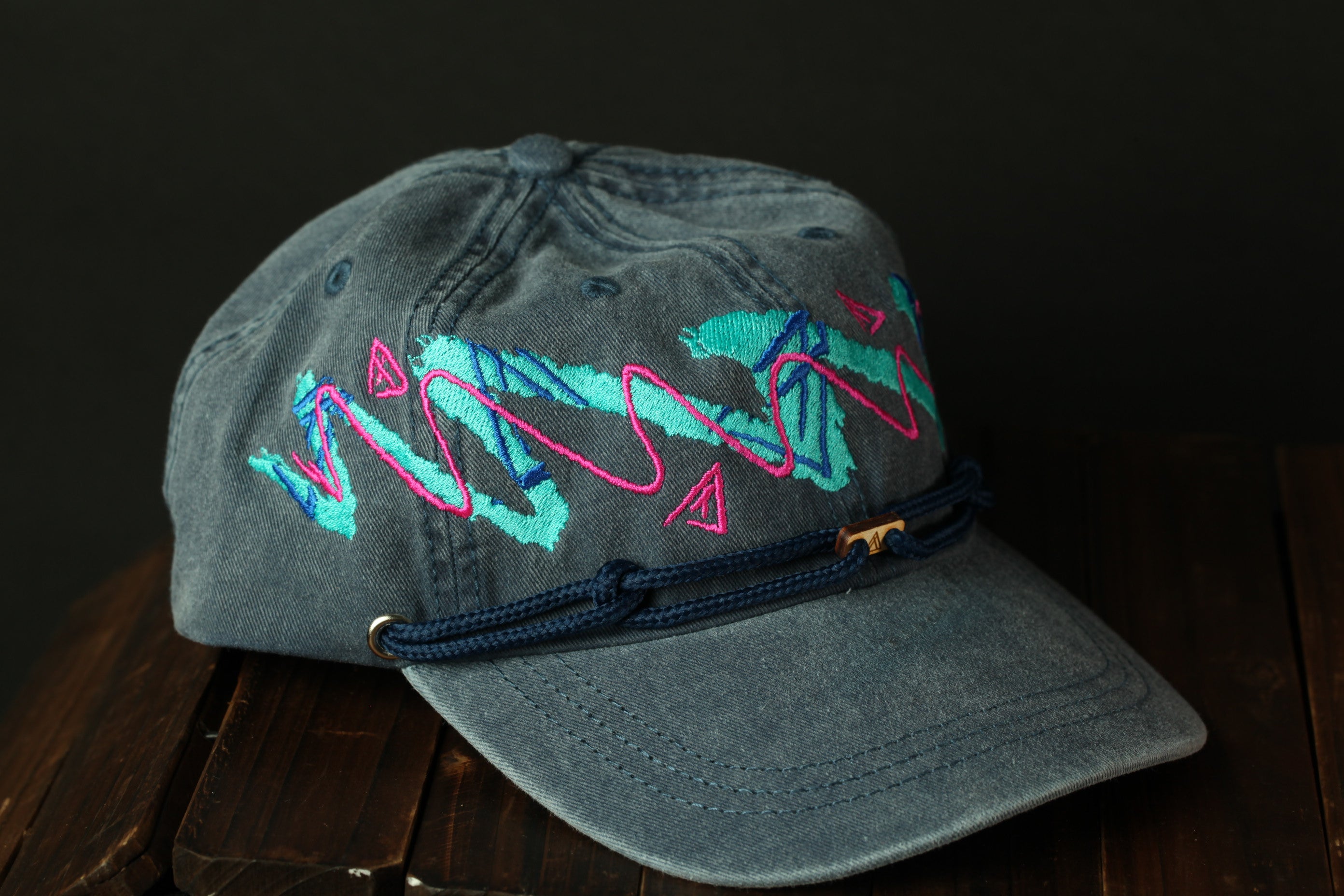 A gray baseball cap with a colorful, abstract embroidered design on the front, resting on a wooden surface.