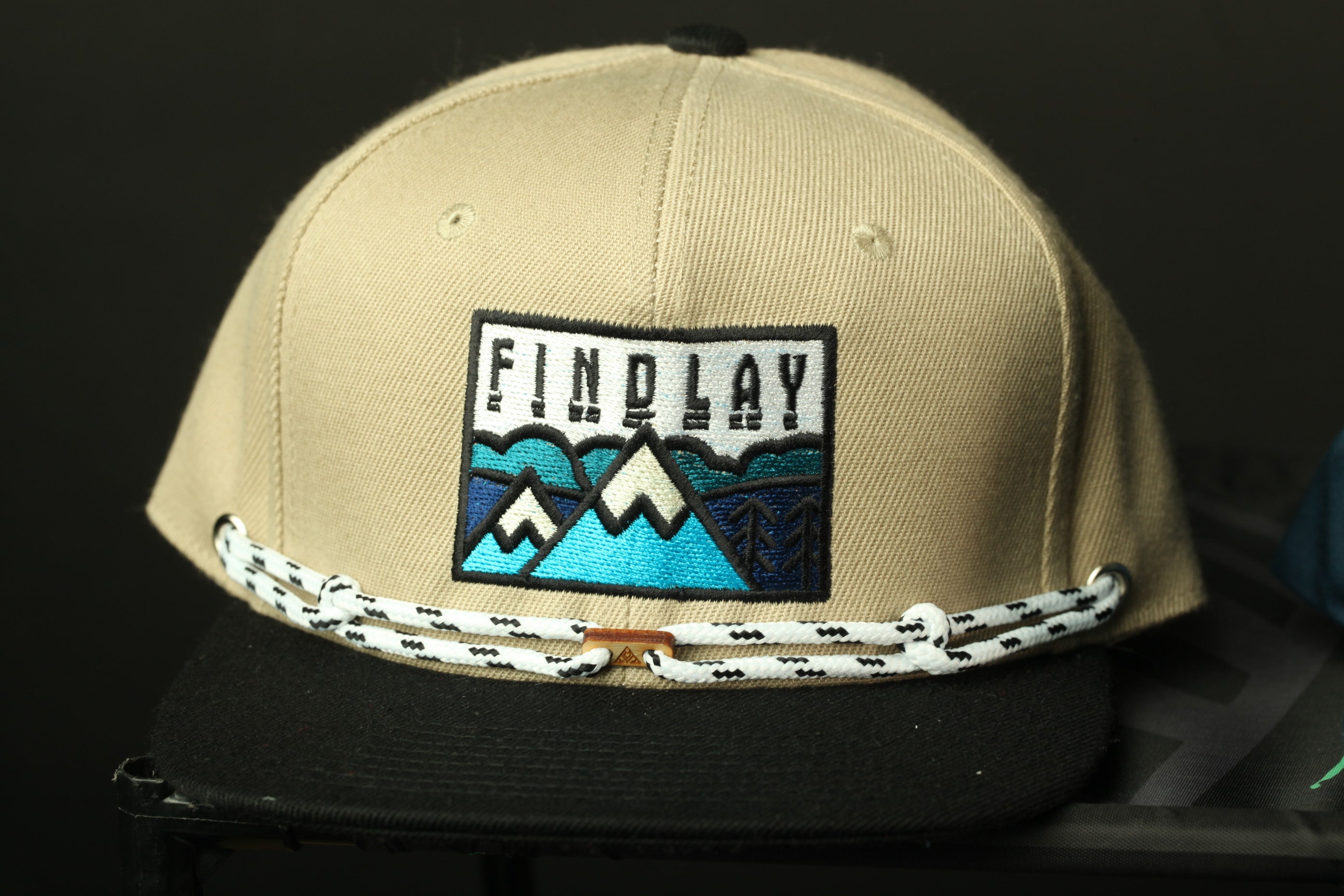 A tan baseball cap with a black brim and a beige logo on the front, featuring a mountain landscape design.