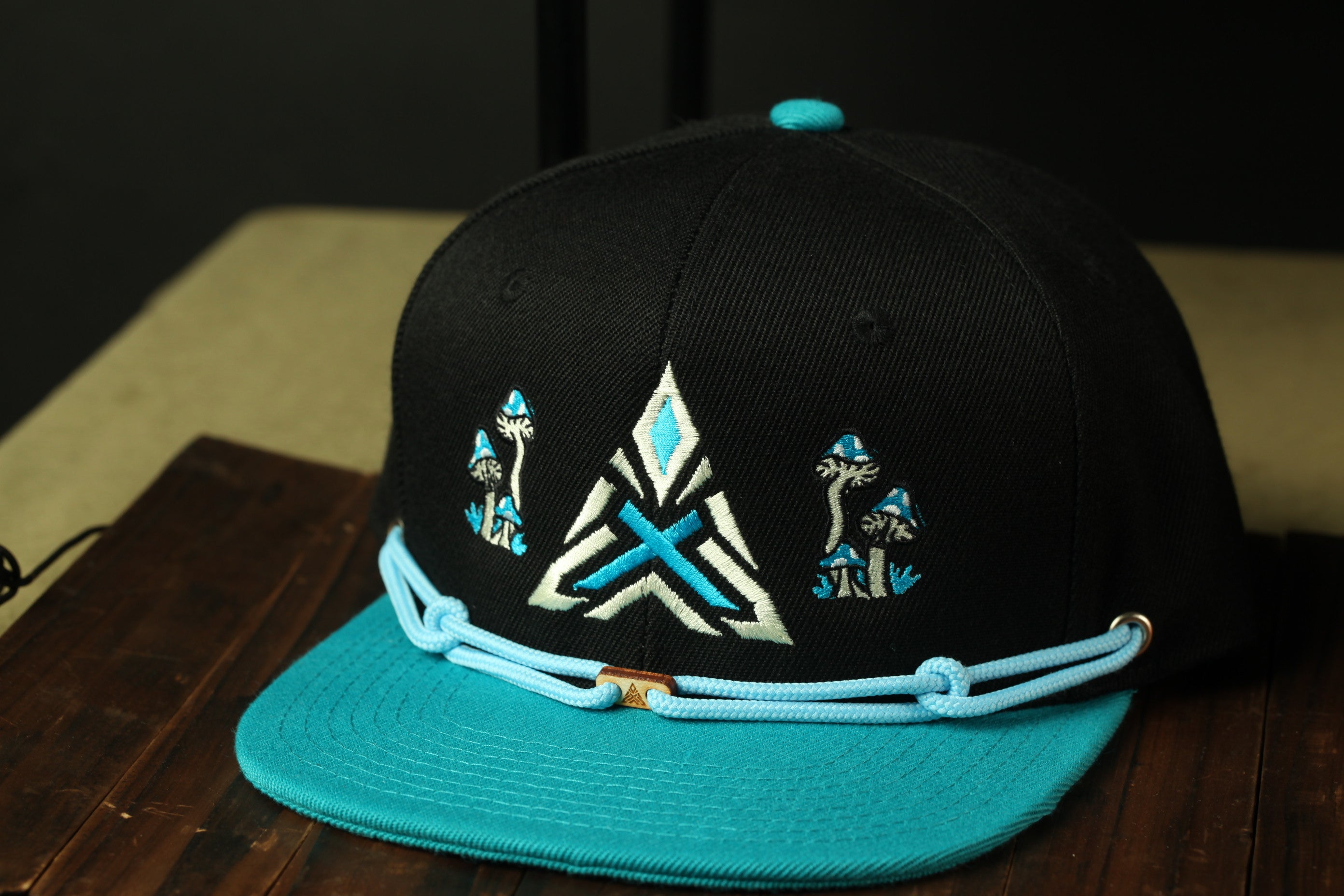 A black baseball cap with a teal brim and a blue and white geometric design on the front, sitting on a wooden surface.