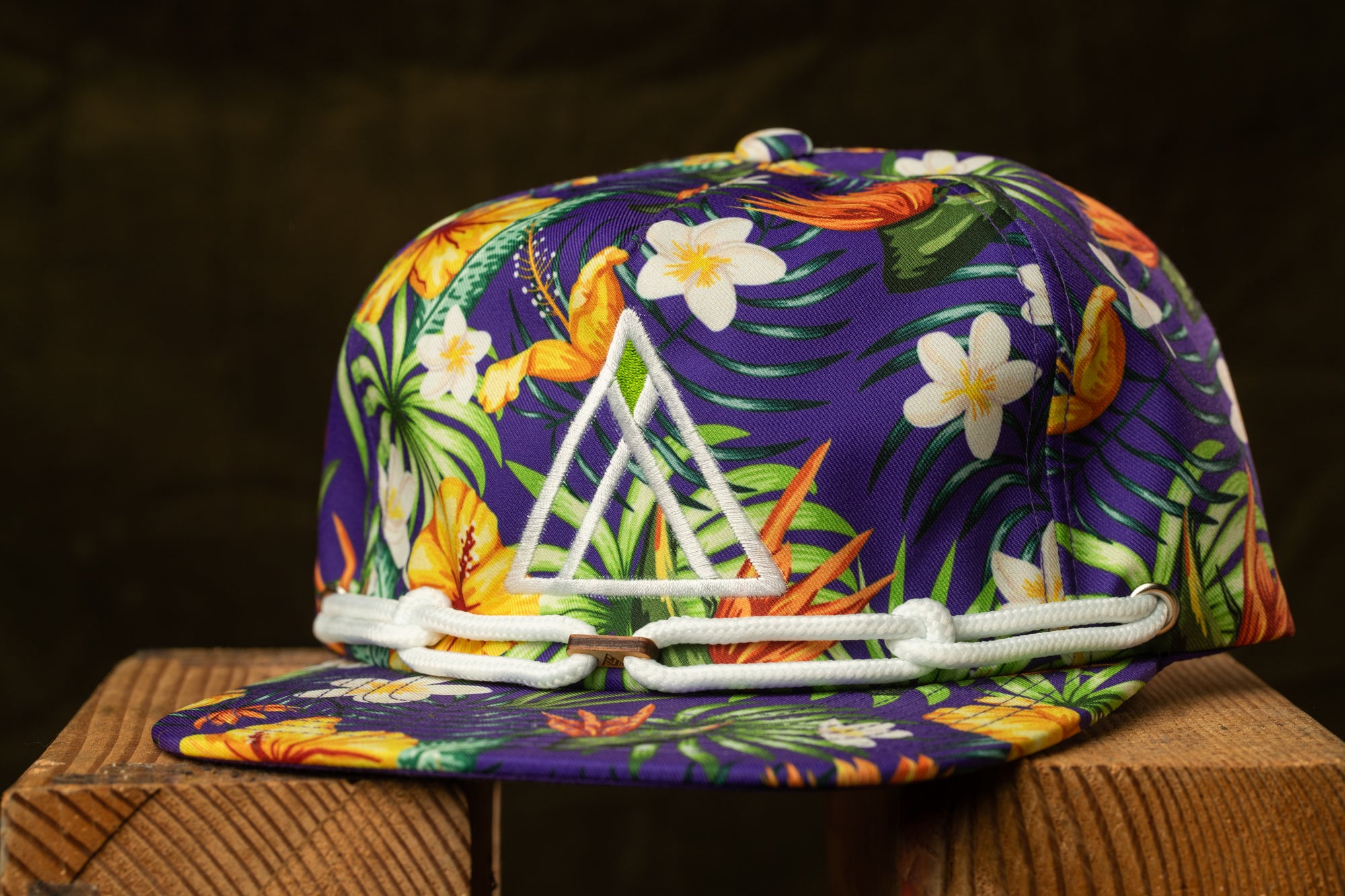 A colorful floral baseball cap with a white triangle logo on the front, displayed on a wooden surface against a dark background.