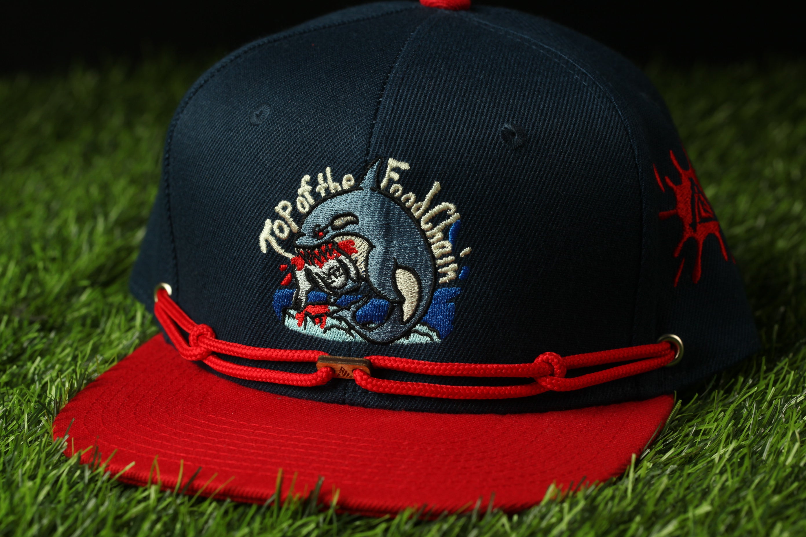A baseball cap with a red brim and a navy blue brim, featuring a graphic of a whale and the text "Top of the Footchain" on the front.