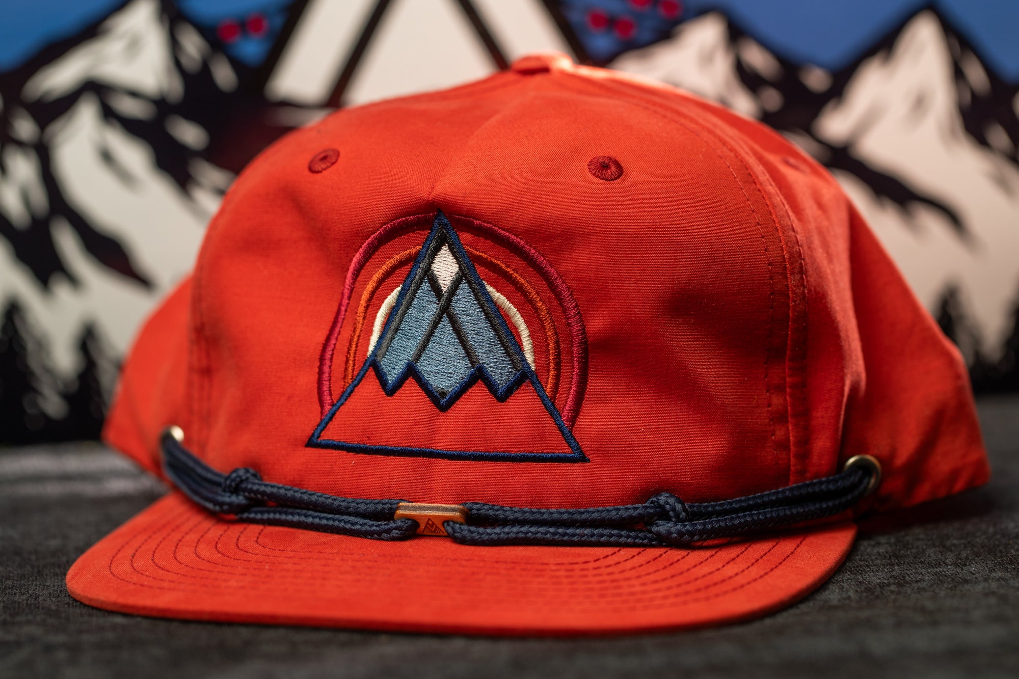 A red baseball cap with a blue and orange geometric design on the front, placed on a gray surface with a mountain landscape in the background.