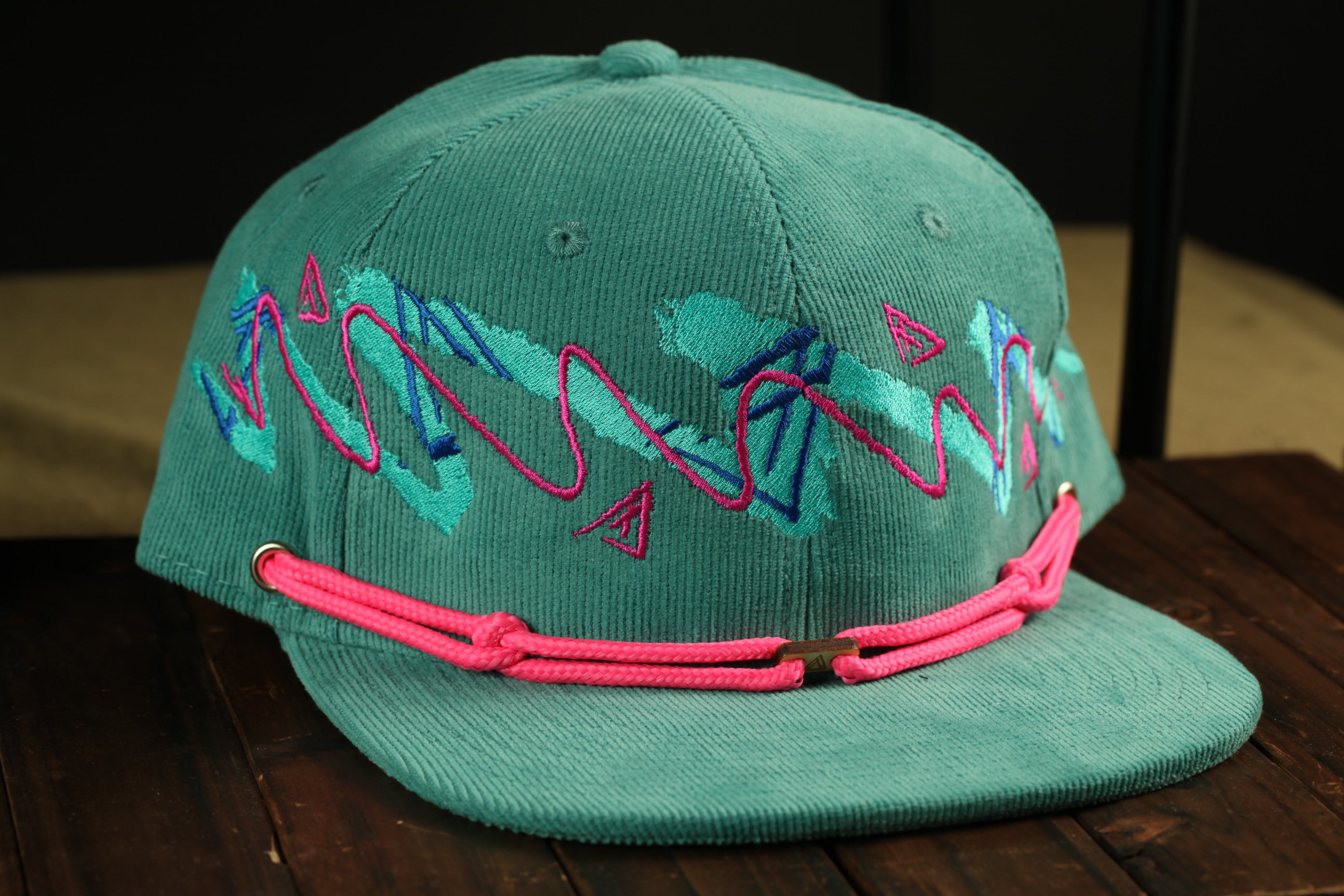 A green baseball cap with pink and blue embroidered designs on it, sitting on a wooden surface.