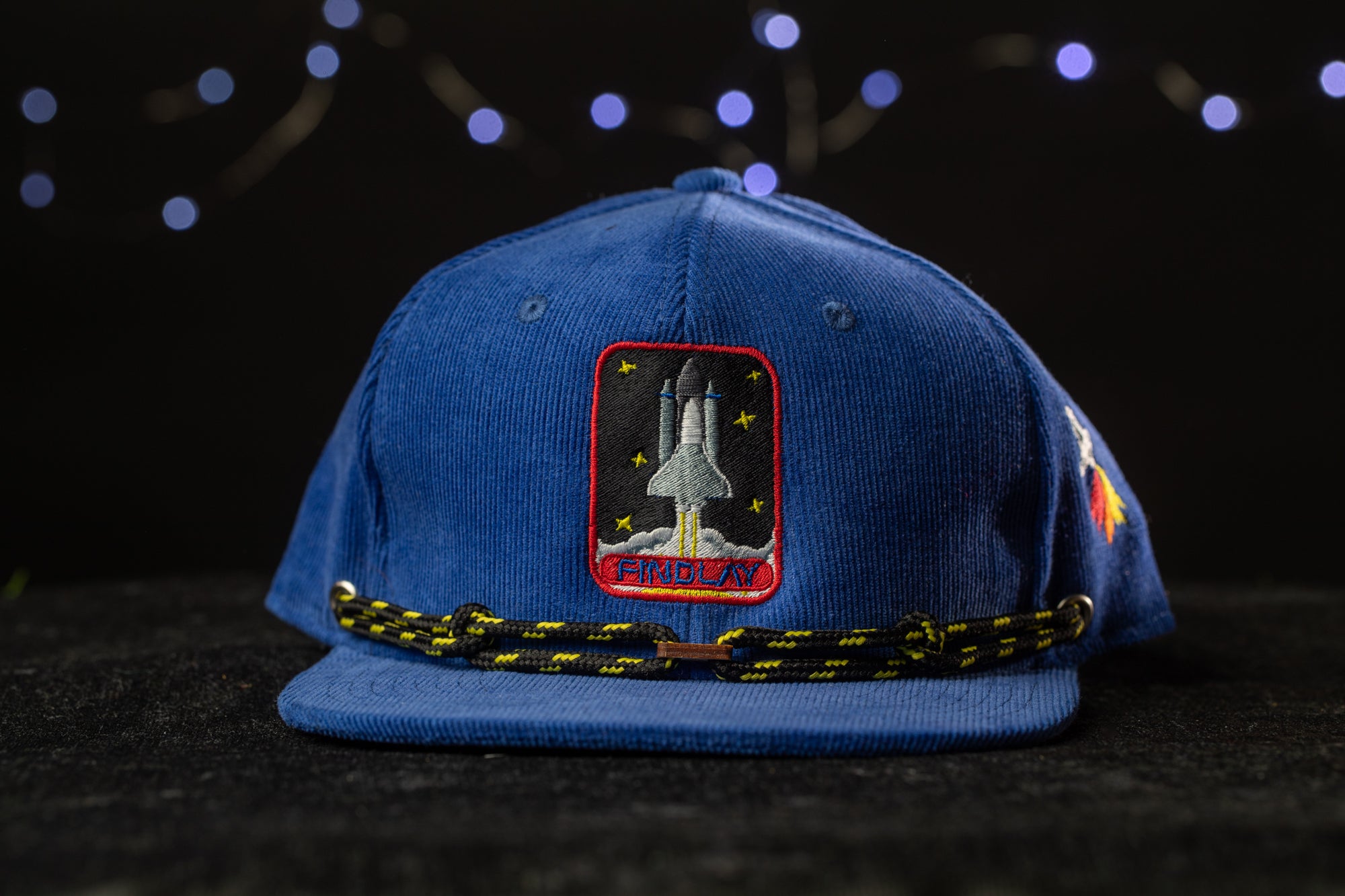 A blue baseball cap with a space shuttle patch on it, placed on a dark surface against a background of twinkling lights.