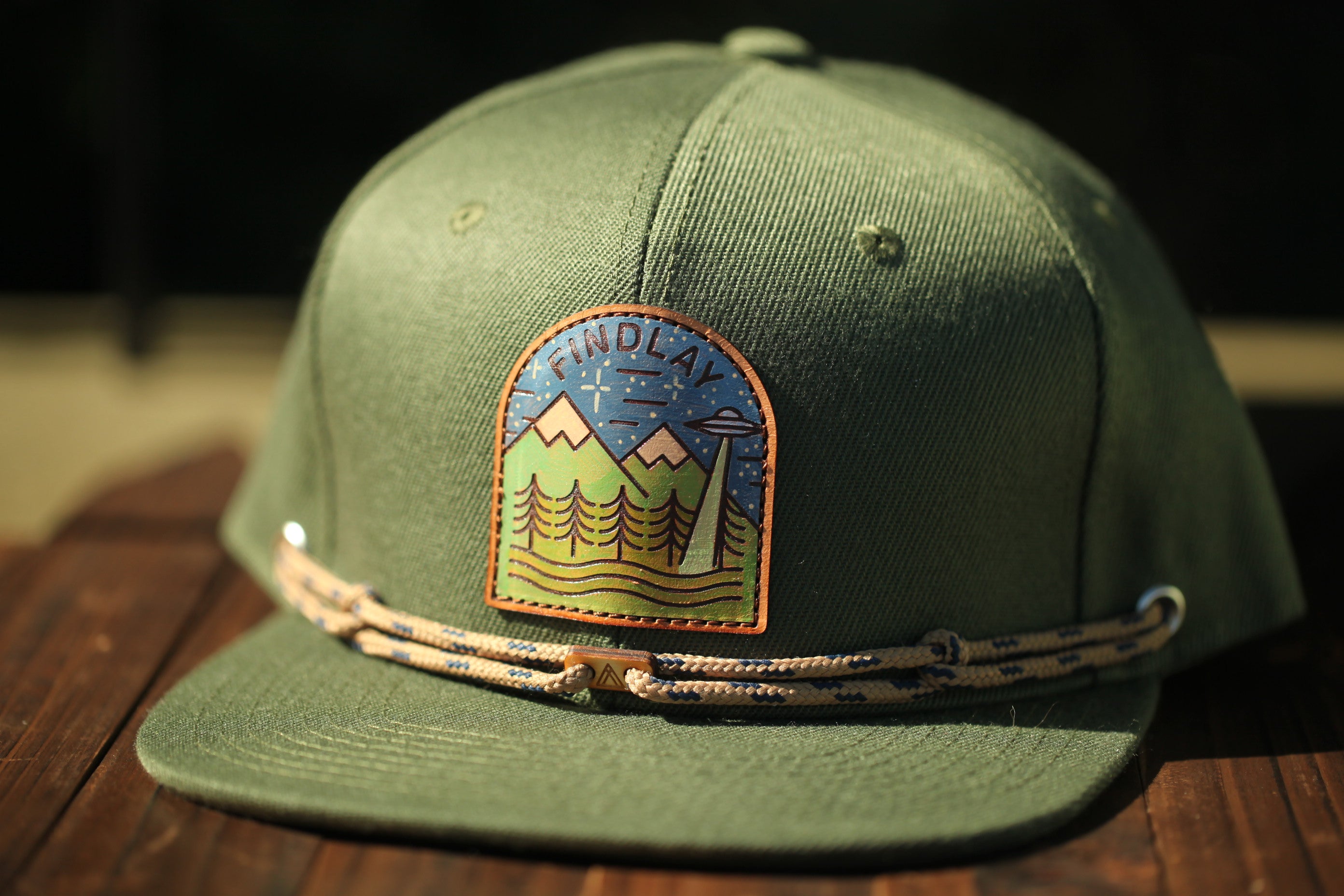 A green baseball cap with a patch featuring a mountain landscape and an alien spacecraft design.