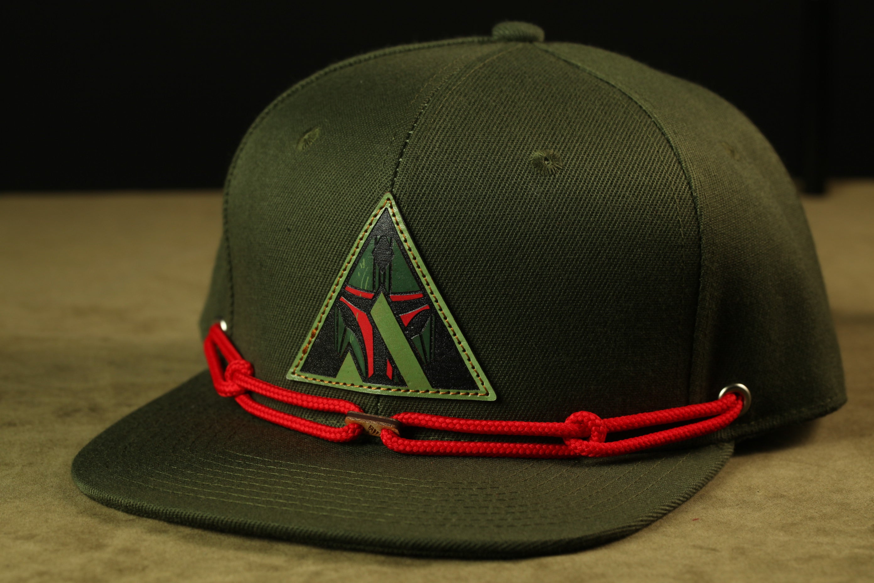 A green baseball cap with a red rope strap and a triangular patch on the front.