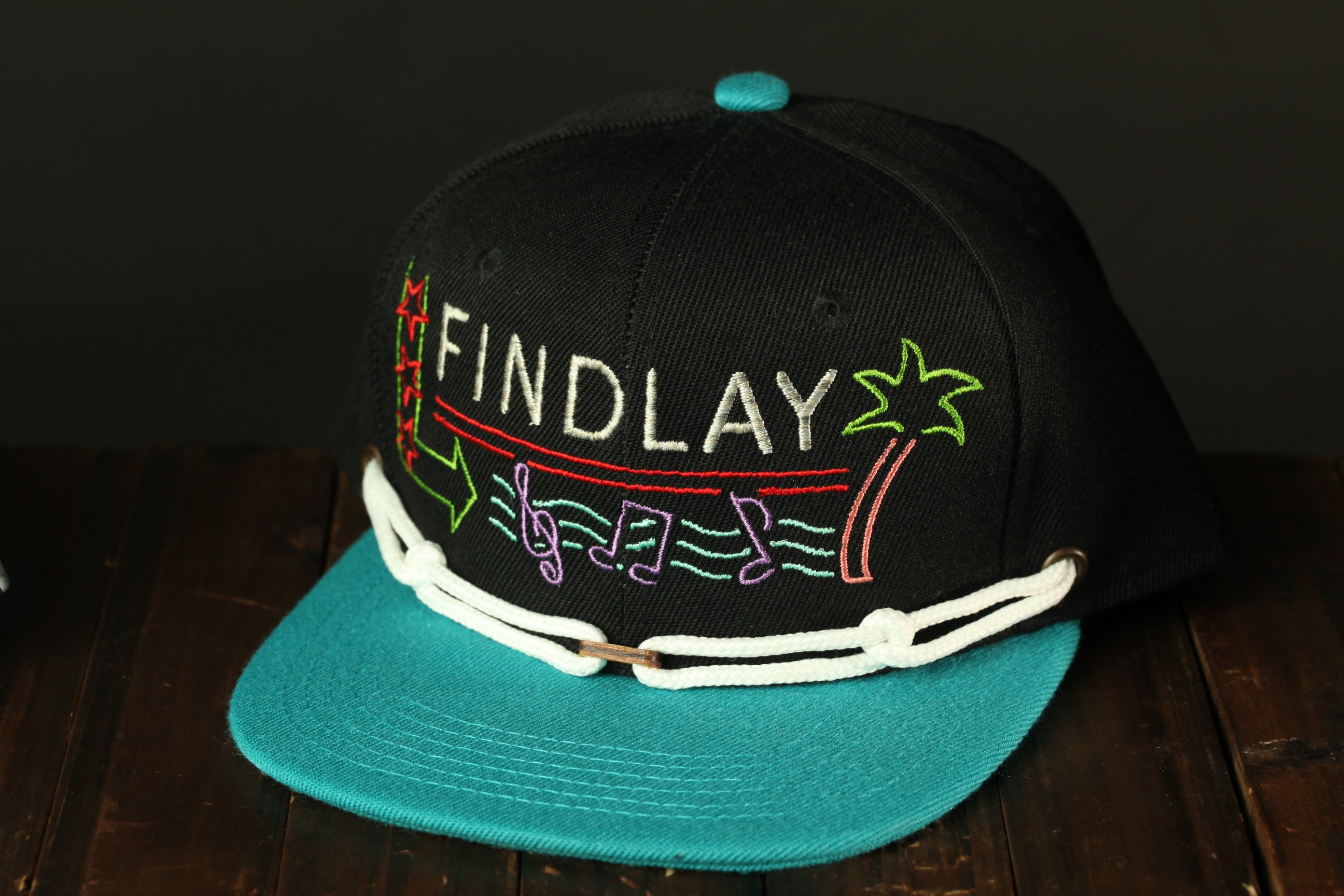 A black baseball cap with colorful embroidered text and graphics, including the word "FINDlay" and a palm tree design.