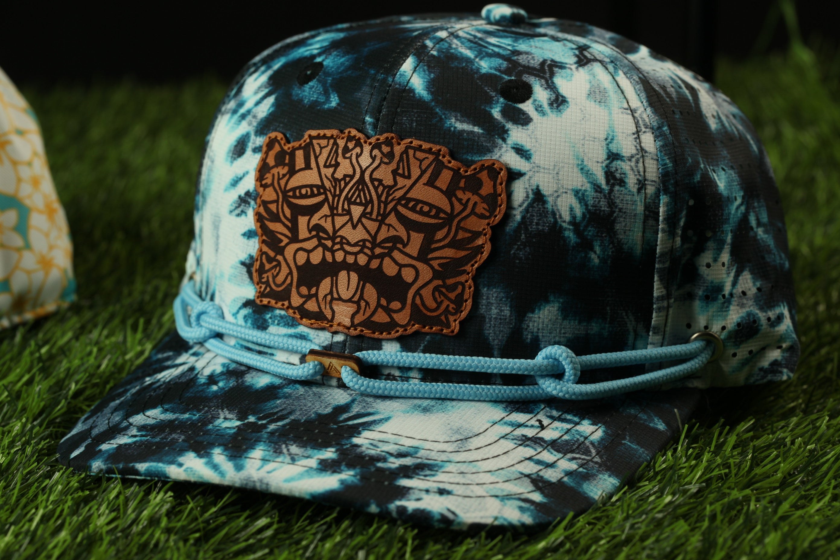 A tie-dye baseball cap with a brown leather patch featuring a stylized face design, resting on a grassy surface.