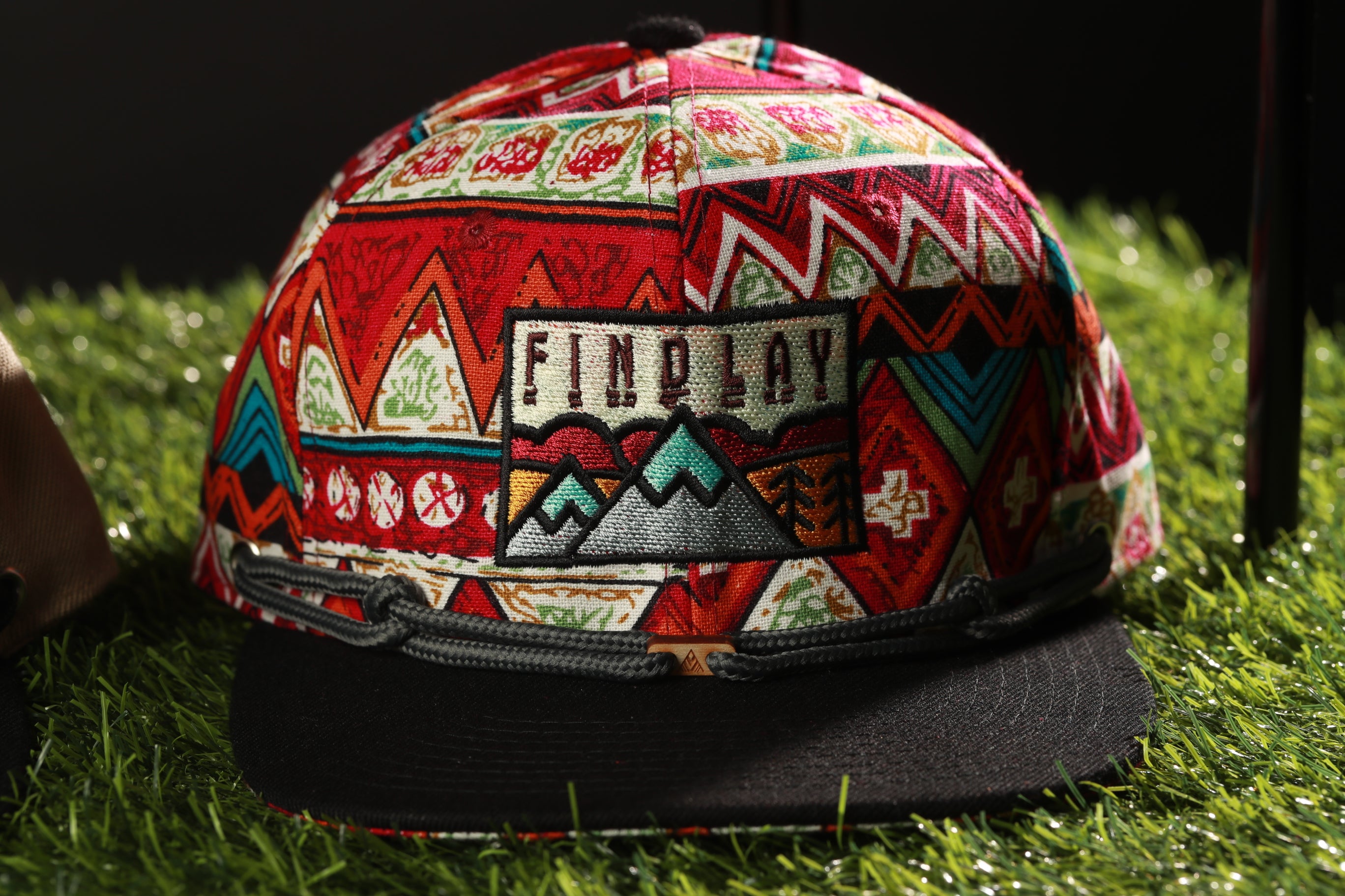 A colorful, patterned baseball cap with a mountain design and the word "FINPLAY" printed on it, resting on a grassy surface.