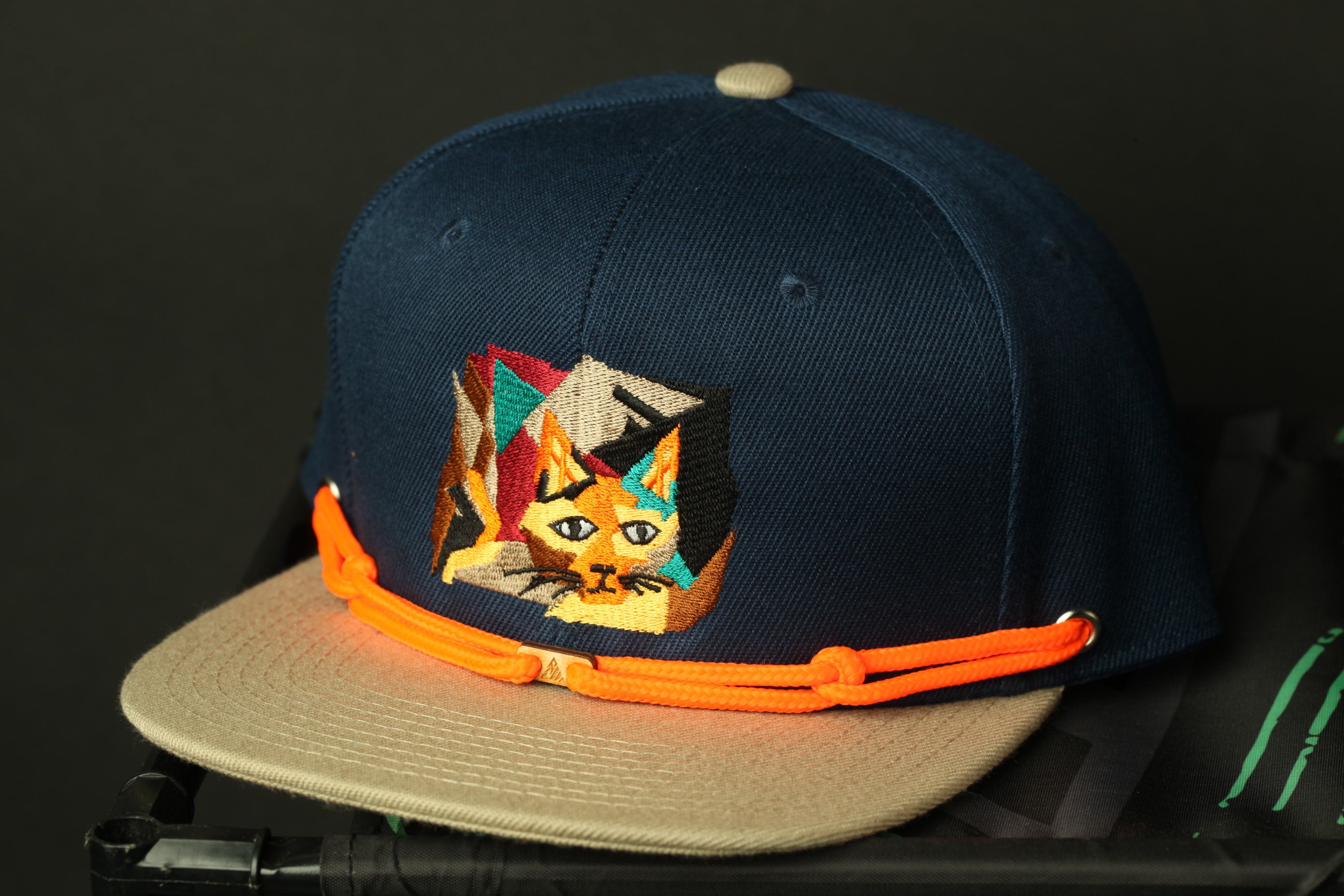 A navy blue baseball cap with a colorful cat embroidered on it, featuring a beige brim and an orange strap.