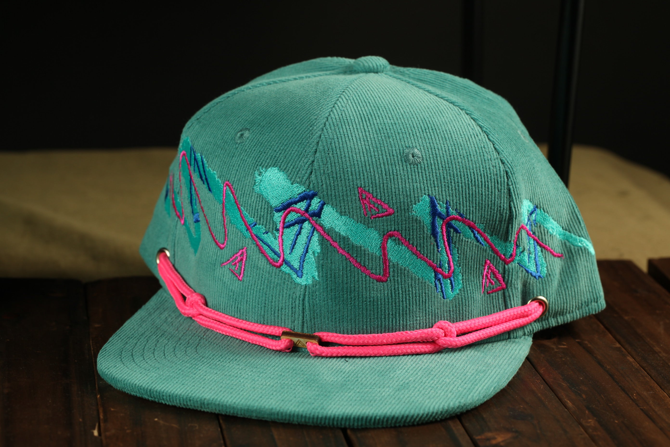 A turquoise baseball cap with pink and blue embroidered designs, featuring a pink adjustable strap.
