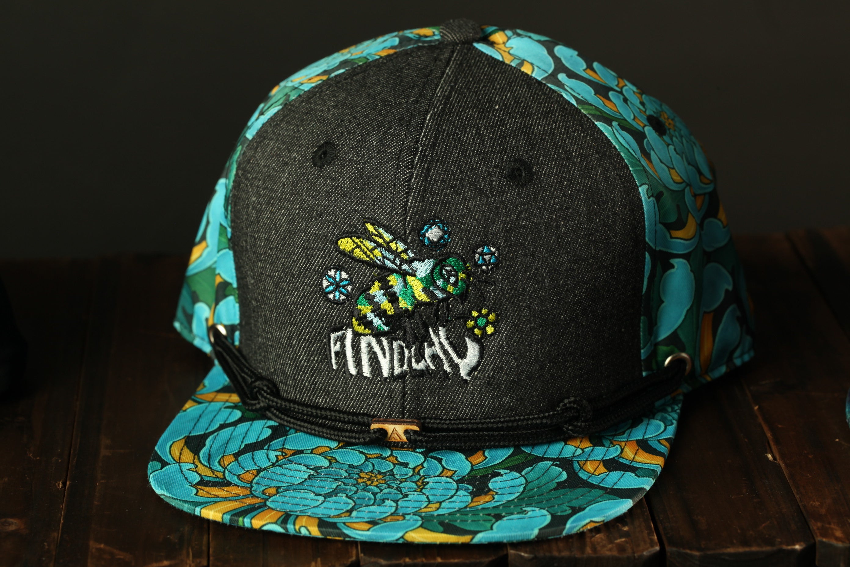 A black baseball cap with a colorful floral pattern and a bee embroidered on it, sitting on a wooden surface.