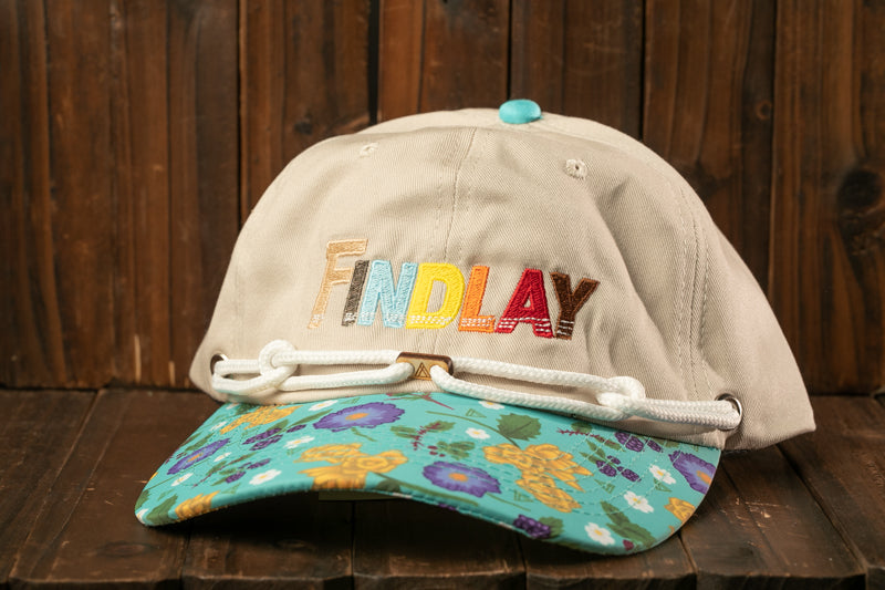 A beige baseball cap with a floral pattern and the word "FINDlay" embroidered on it, placed on a wooden surface.