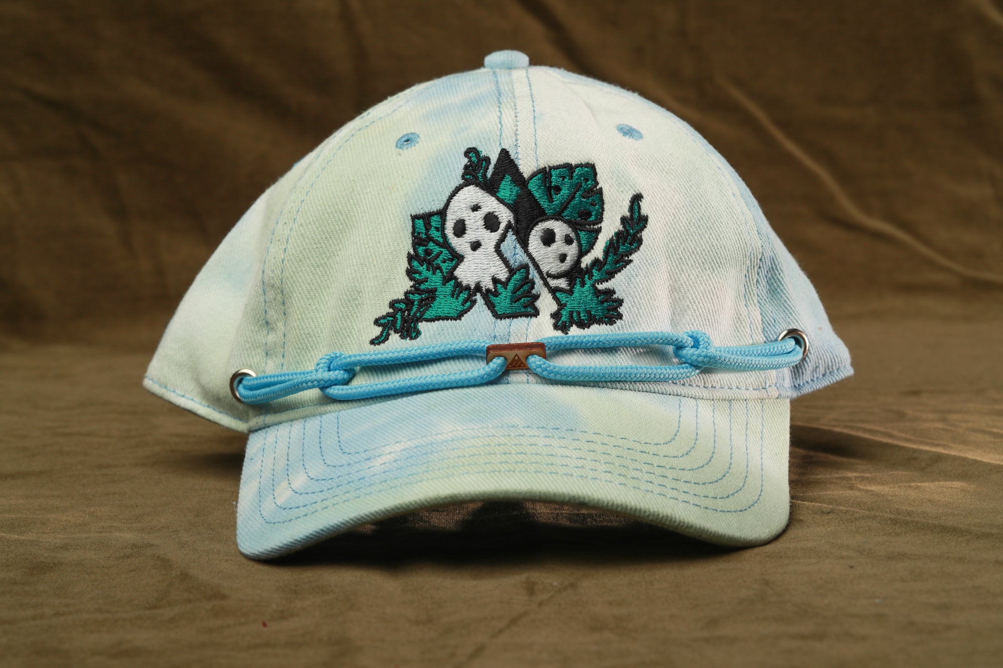 A light blue baseball cap with a unique embroidered design featuring two ghostly figures and green foliage.