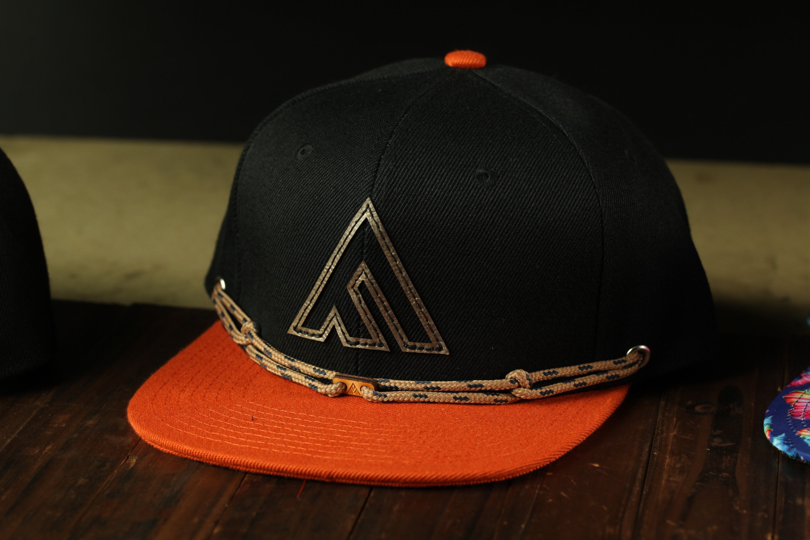 A black baseball cap with an orange brim and a gold-colored logo on the front, sitting on a wooden surface.