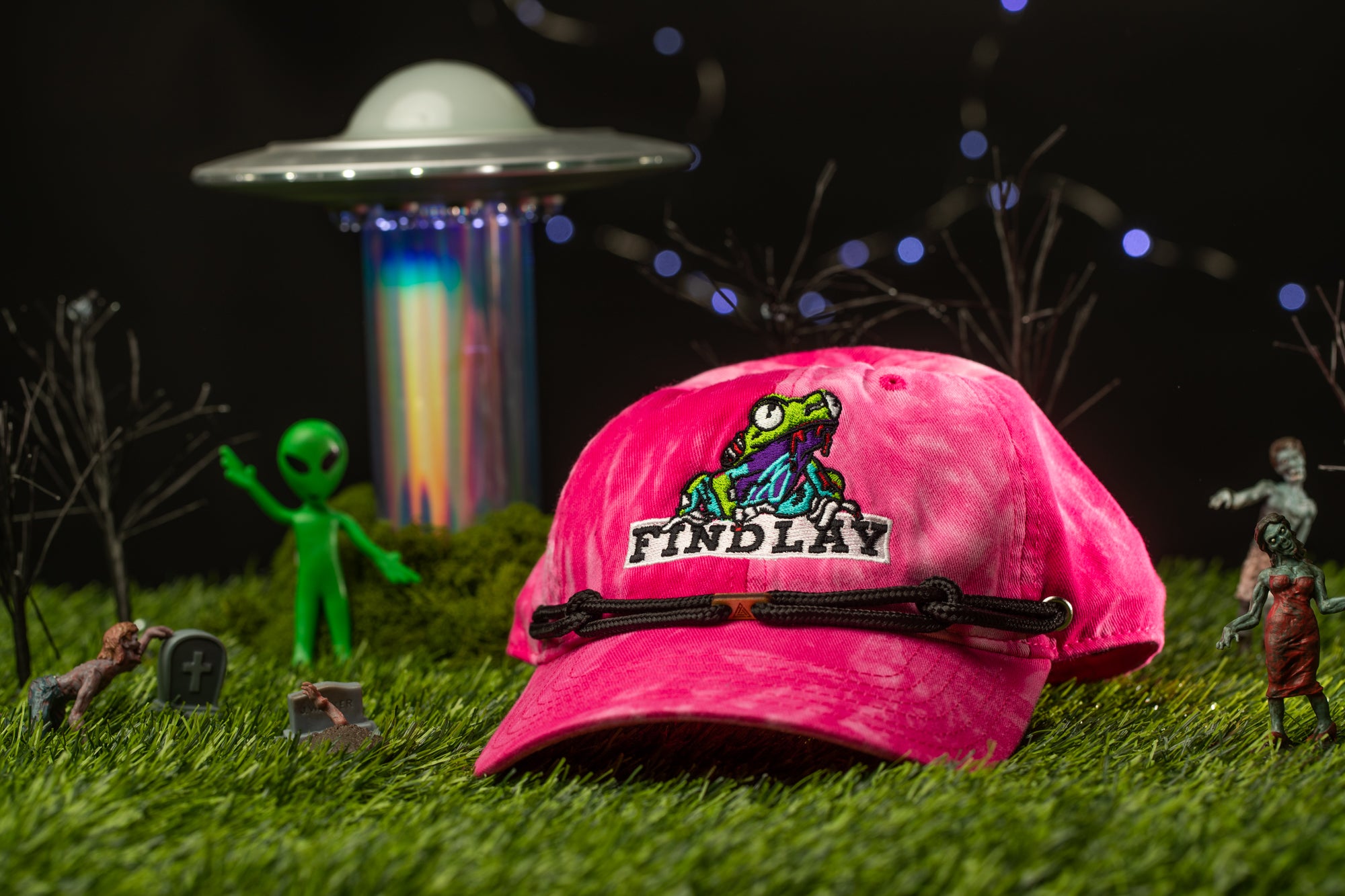 a pink baseball cap with a cartoon frog design, surrounded by various objects such as a green alien figure, a skull, and a spaceship.