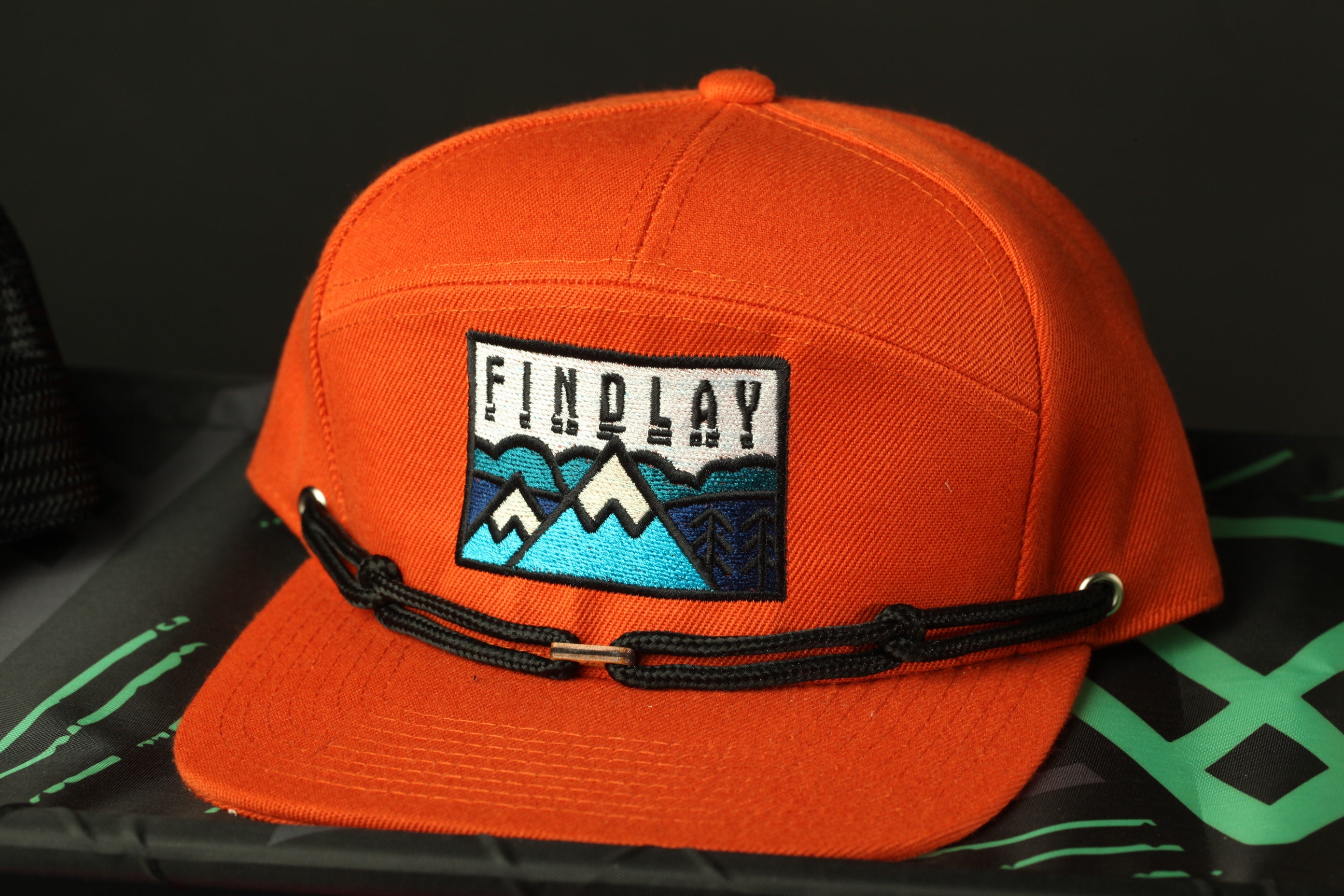 An orange baseball cap with a black strap and a patch on the front, featuring a mountain landscape design.