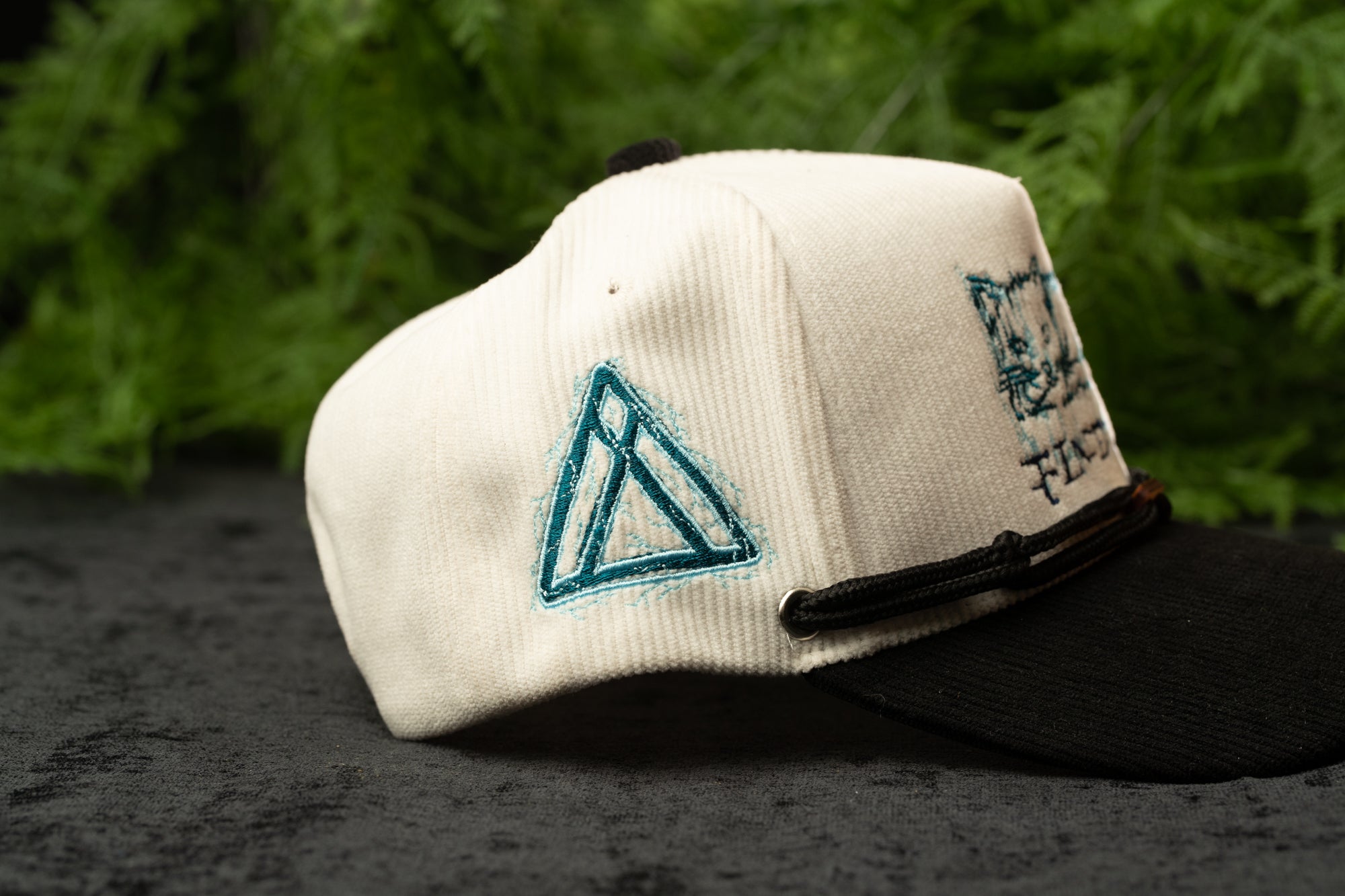 A white baseball cap with a black brim, featuring a blue triangle design on the front, is shown against a blurred green background.