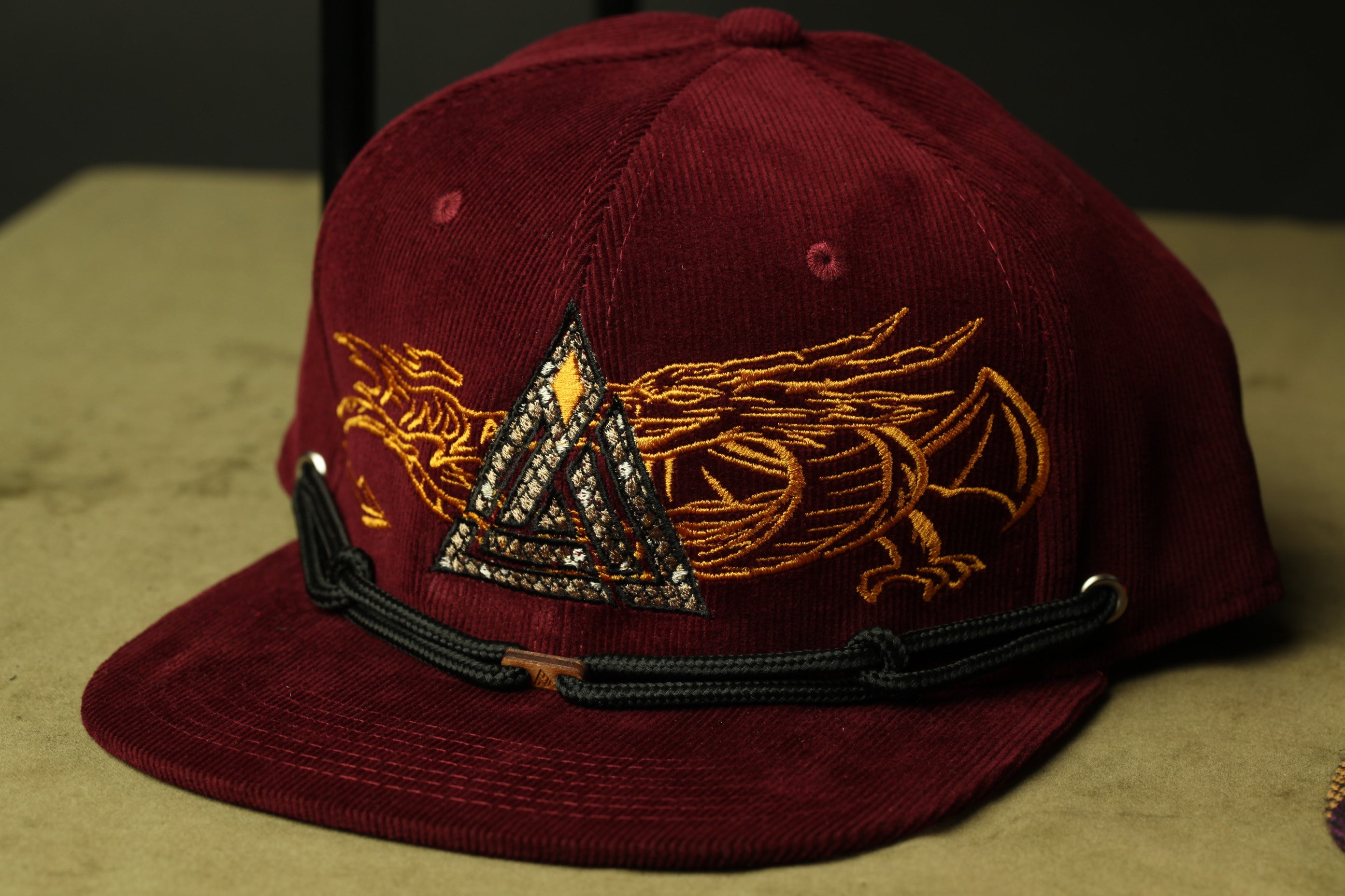 A burgundy baseball cap with a gold dragon design and a triangular emblem on the front.