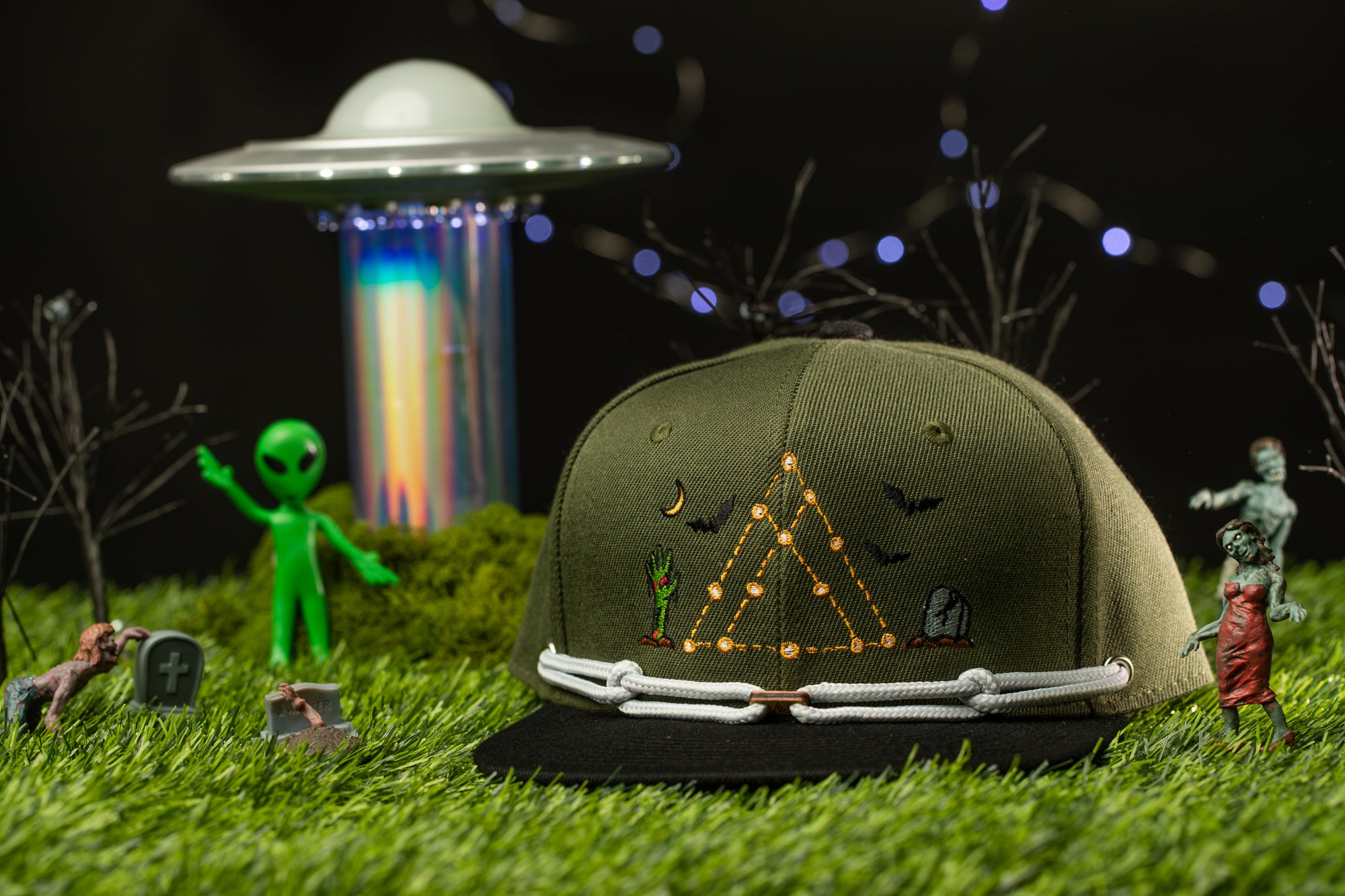 a green baseball cap with various alien and celestial motifs, surrounded by small toy figures and a spaceship in a dark, starry background.