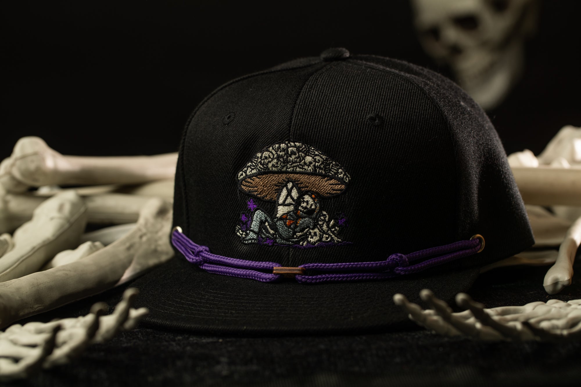 A black baseball cap with a mushroom design and purple string is displayed on a surface with white bones.