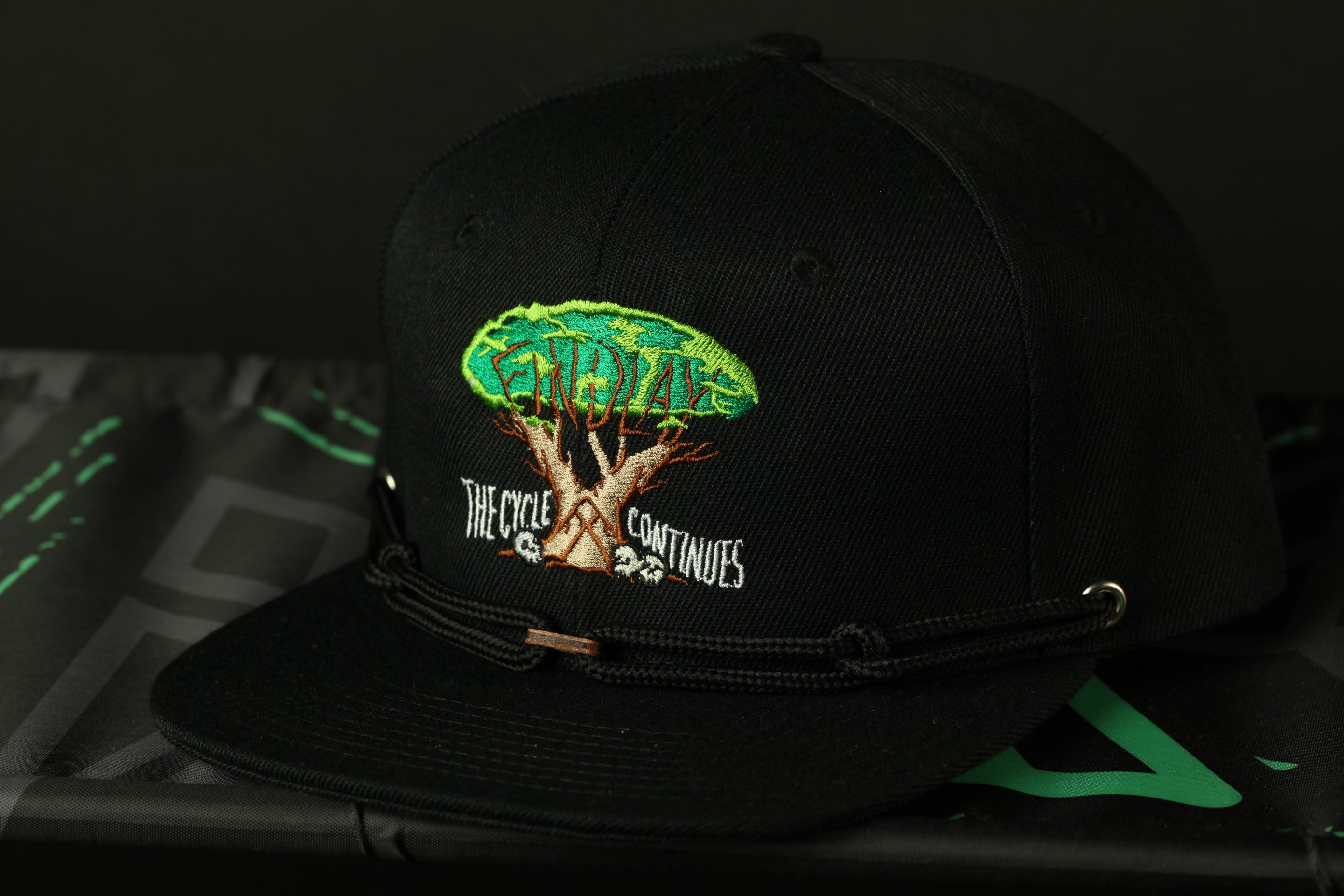 A black baseball cap with a tree design and text on it, placed on a surface with a green and black patterned background.