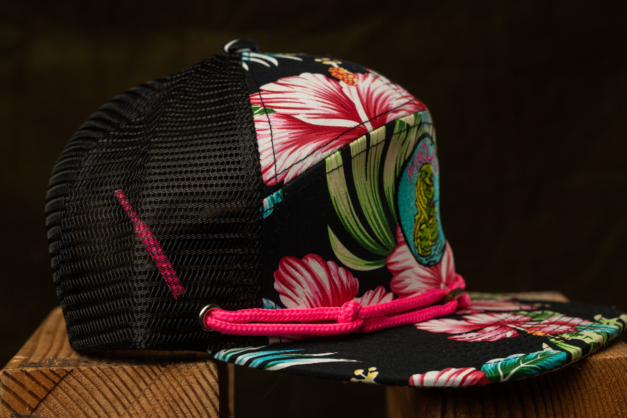 A colorful floral-patterned baseball cap with a black mesh panel and a pink braided strap, resting on a wooden surface.