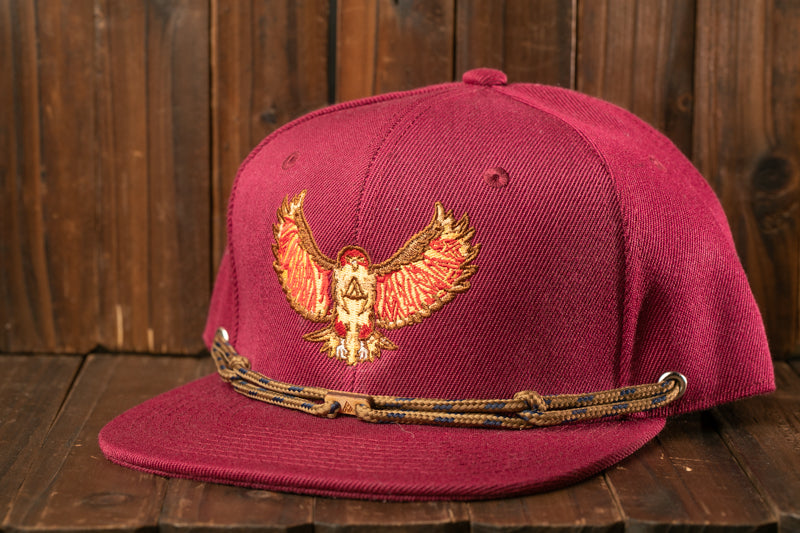 A maroon baseball cap with a red and gold embroidered eagle design on the front, sitting on a wooden surface.