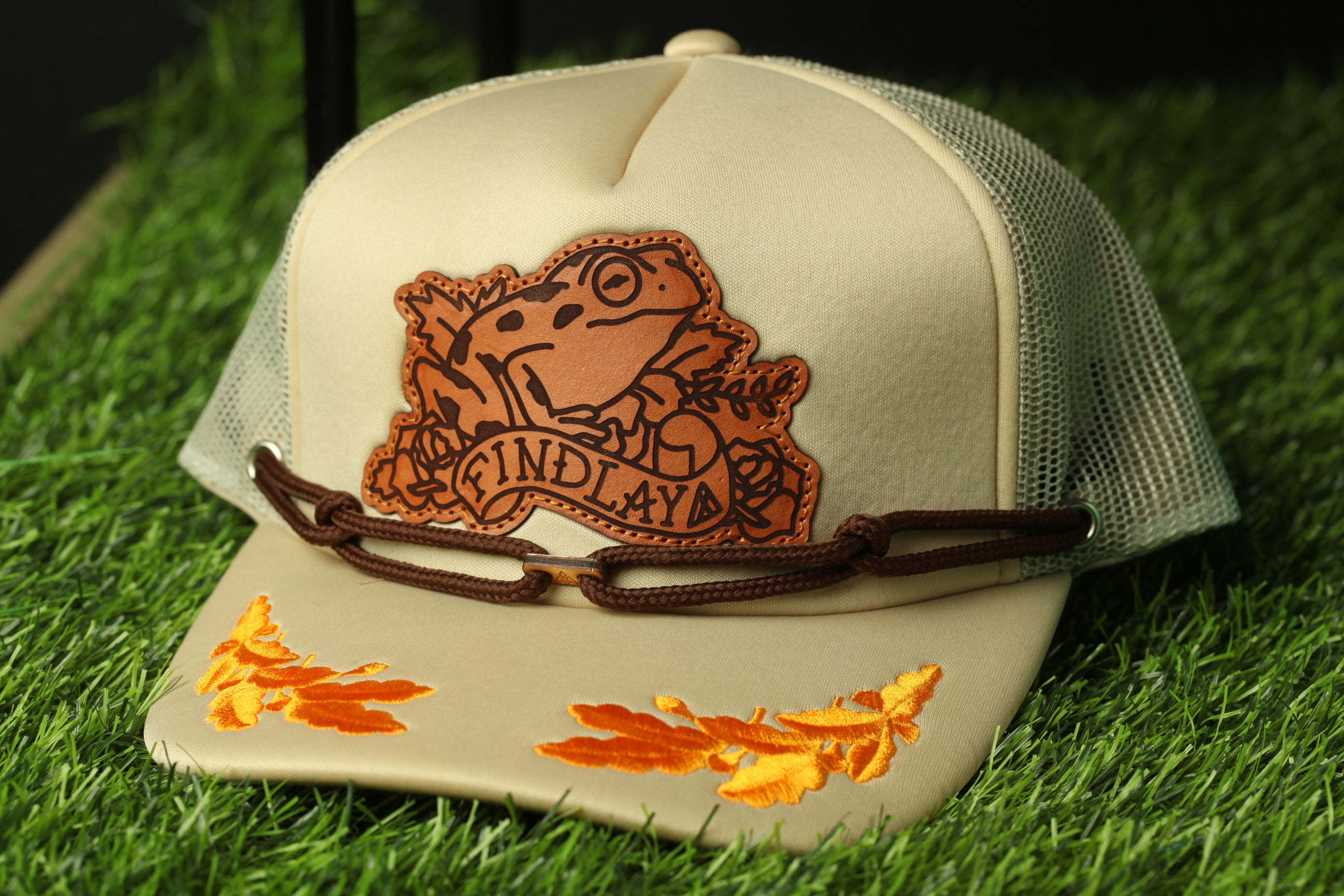 A beige baseball cap with a brown leather patch featuring a frog and the text "FIND LAY" on it, placed on a grassy surface.