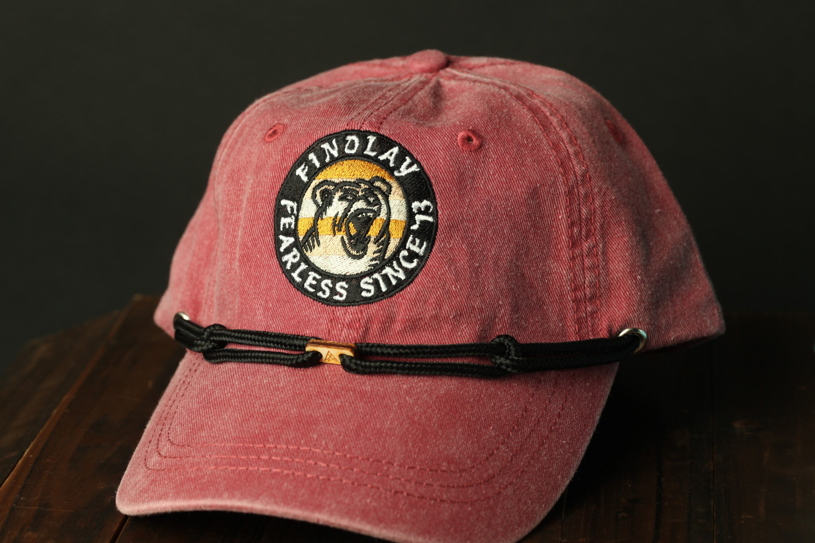 A red baseball cap with a black and gold logo on it, featuring the text "FINDOLAY FEARLESS SINCE" and a bear image.