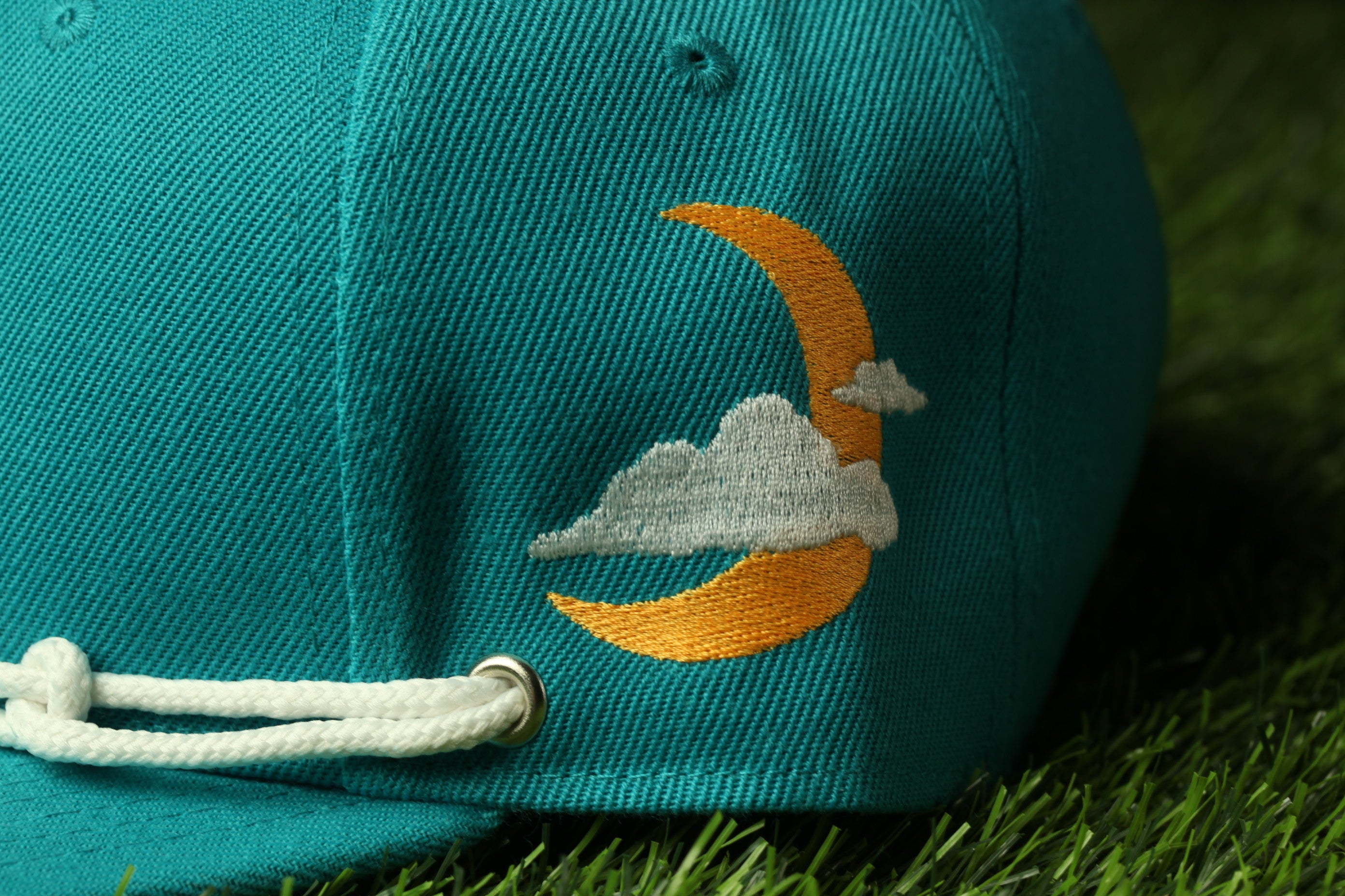 A close-up view of a teal baseball cap with an embroidered design of a crescent moon and clouds on the side.