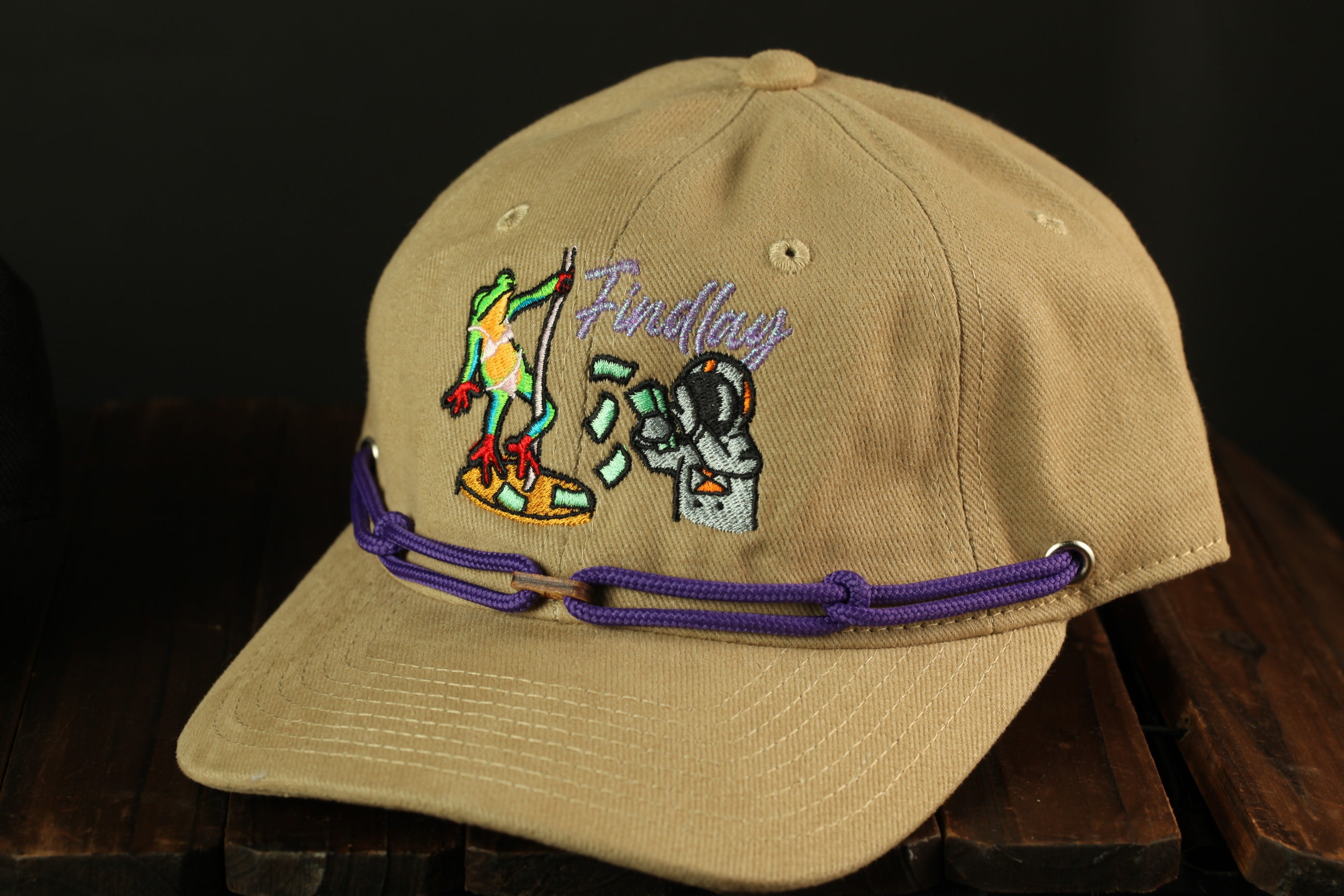A beige baseball cap with a purple string and a cartoon frog on it, sitting on a wooden surface.