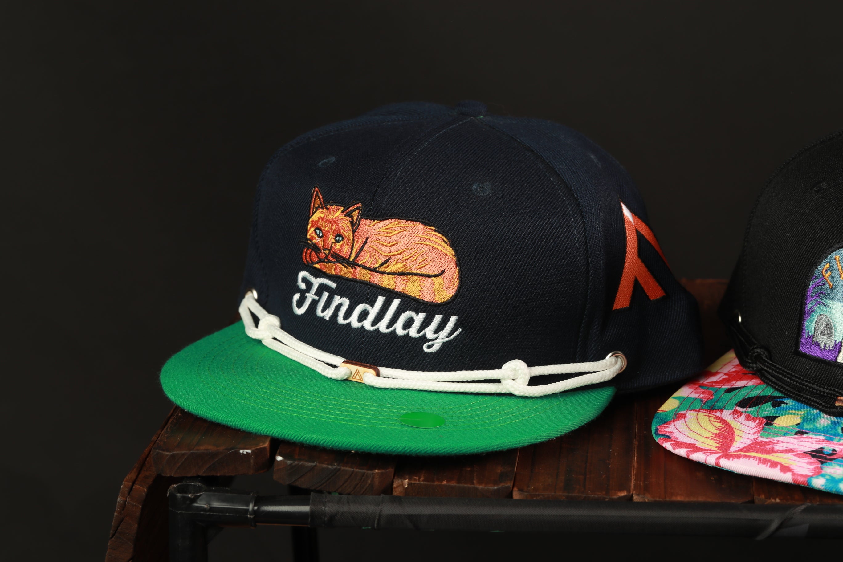 A black baseball cap with a green brim and a cat embroidered on it, sitting on a wooden surface.