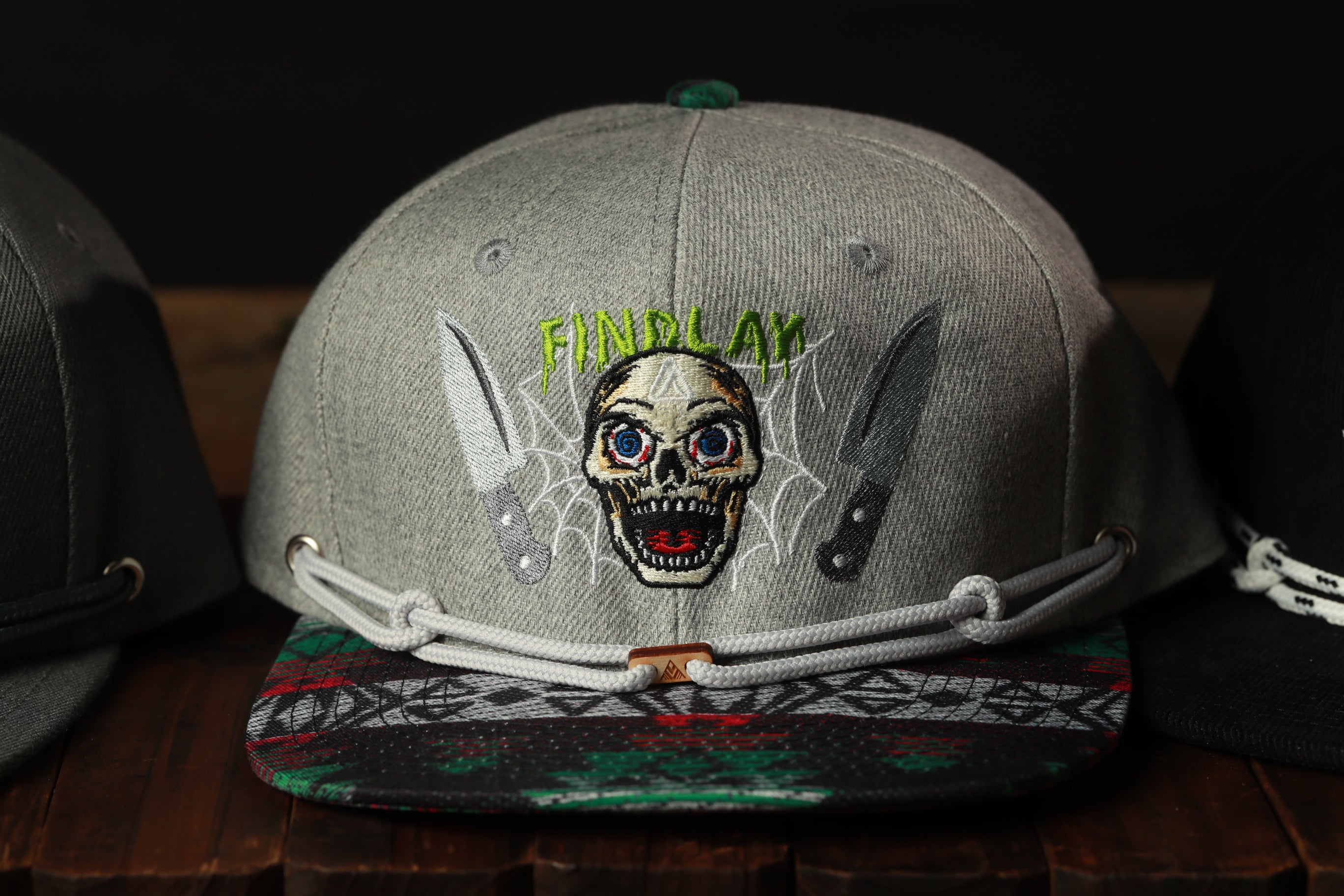 A gray baseball cap with a skull design and the text "FINPLAY" on it, along with two knives, is displayed on a wooden surface.